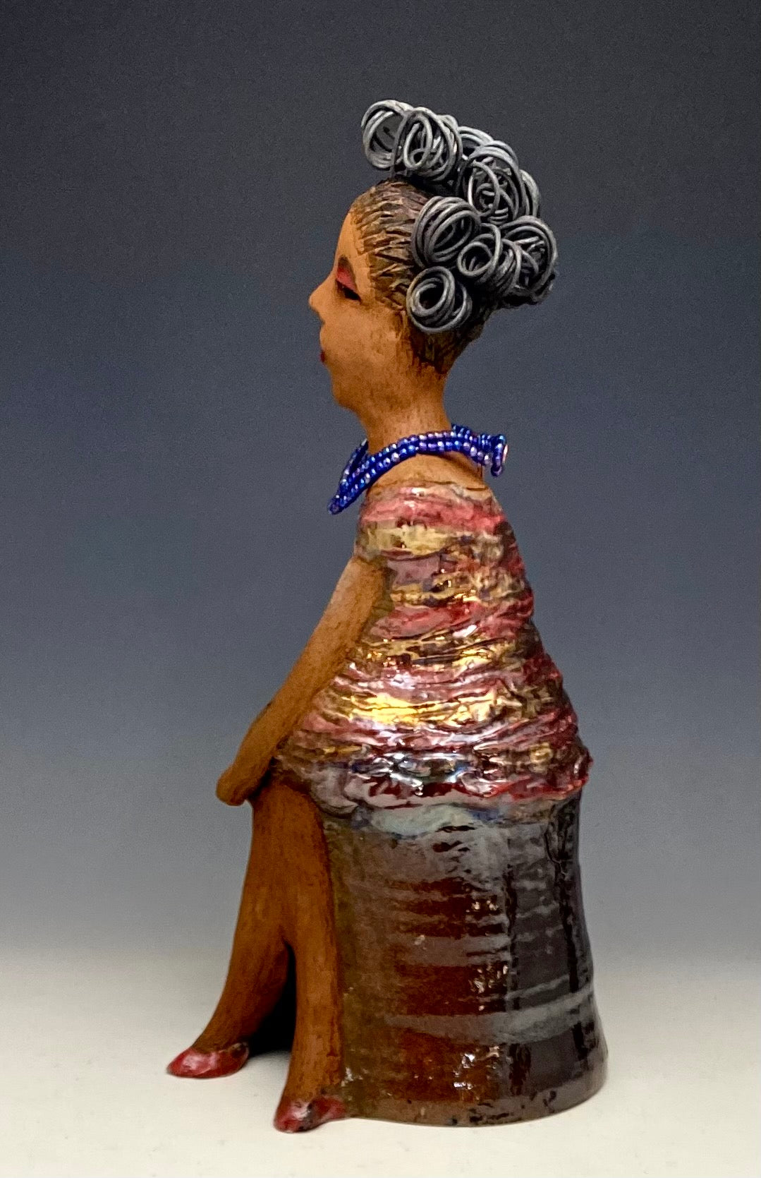 You know someone like Kim. One who reaps elegance and sophistication! Kim stands 13" x 5" x 7" and weighs 2.07 lbs. She has a lovely light beige complexion with over 4 feet of 16 gauge spiral coiled wire hair. Kim's metallic copper red and gold dress is accented by a multicolored aqua blue beaded necklace. Kim's maroon eye shadow gives her a distinguished mystical look. One must wonder what is Kim thinking? Kim sits with her legs crossed as she awaits a special place to grace your home.