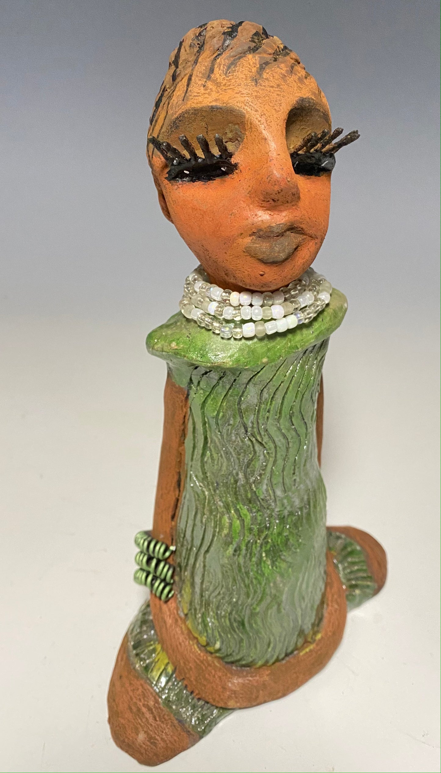 Meet Gracie! GracIe stands 9.5" x 6" x 3.5" and weighs 1.06 lbs. She has a lovely beige brown complexion with long etched braided clay hair. Gracie has a long white and clear beaded necklace. Gracie wears long stylish eyelashes Her long loving arms rest beside her copper green dress. Gracie is a sophisticated lady that will grace your home.