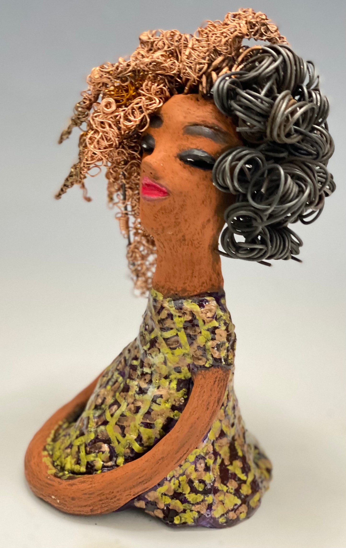 Meet Kalifa, an elegant figurine weighing 1.11 lbs and measuring 8" x 5" x 5.5". Her glossy spotted metallic copper glazed dress and long twisted wire locs are expertly crafted. With her honey brown complexion and red lips, she is sure to capture the attention of your guests. Her daring two-tone copper hair, which took 6 hours to create, adds to her beauty. Invite her into your home and spark meaningful conversations with her cheerful eyes and hopeful gaze.