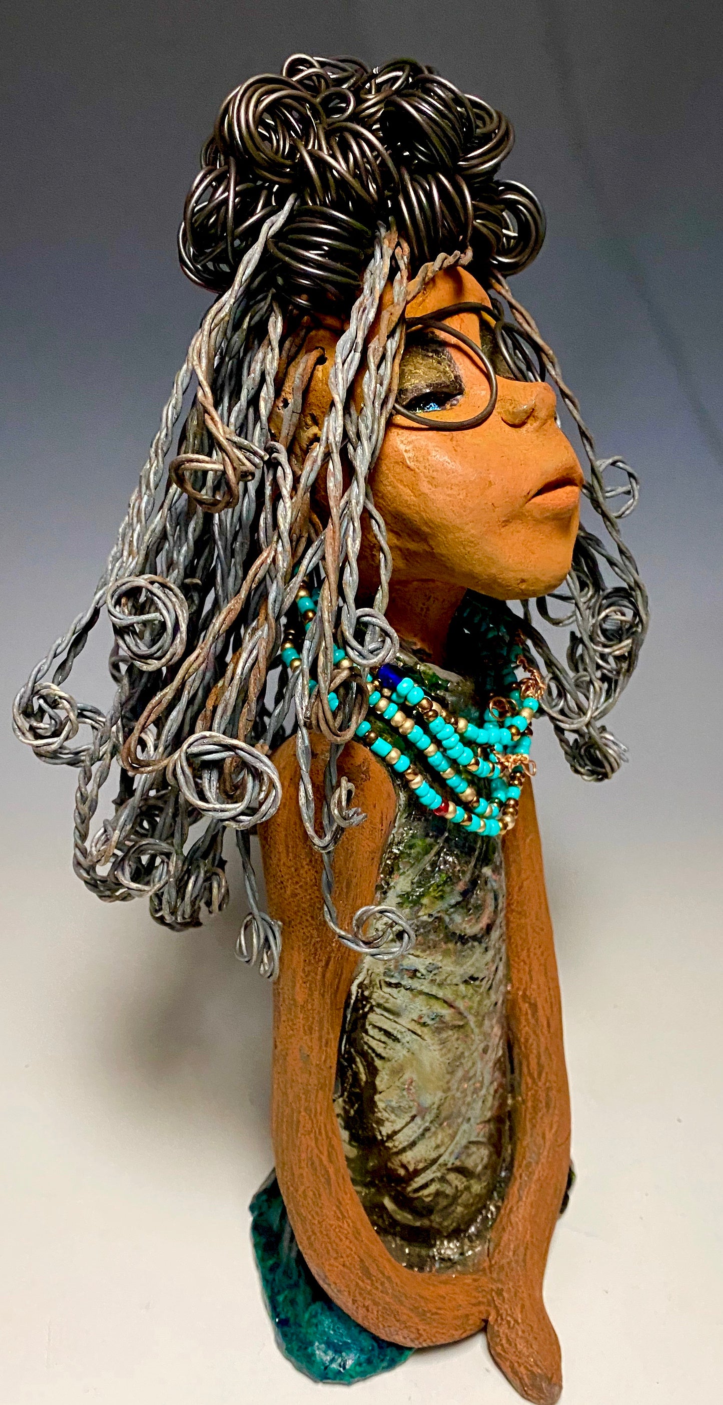 Meet Clarice! Clarice stand 13" x 6" x 5" and weighs 4.03 lbs. She has a lovely honey brown complexion with big bright eyes. Clarice wears glasses now! Her Herdew took over 2 hours to complete. It's made with 16 gauge wire. Clarice's dress is alligator green with streaks of copper. She has a blue beaded spiral necklace around her neck. Clarice looks to have a New ExtrArtindary Vision for 2021!