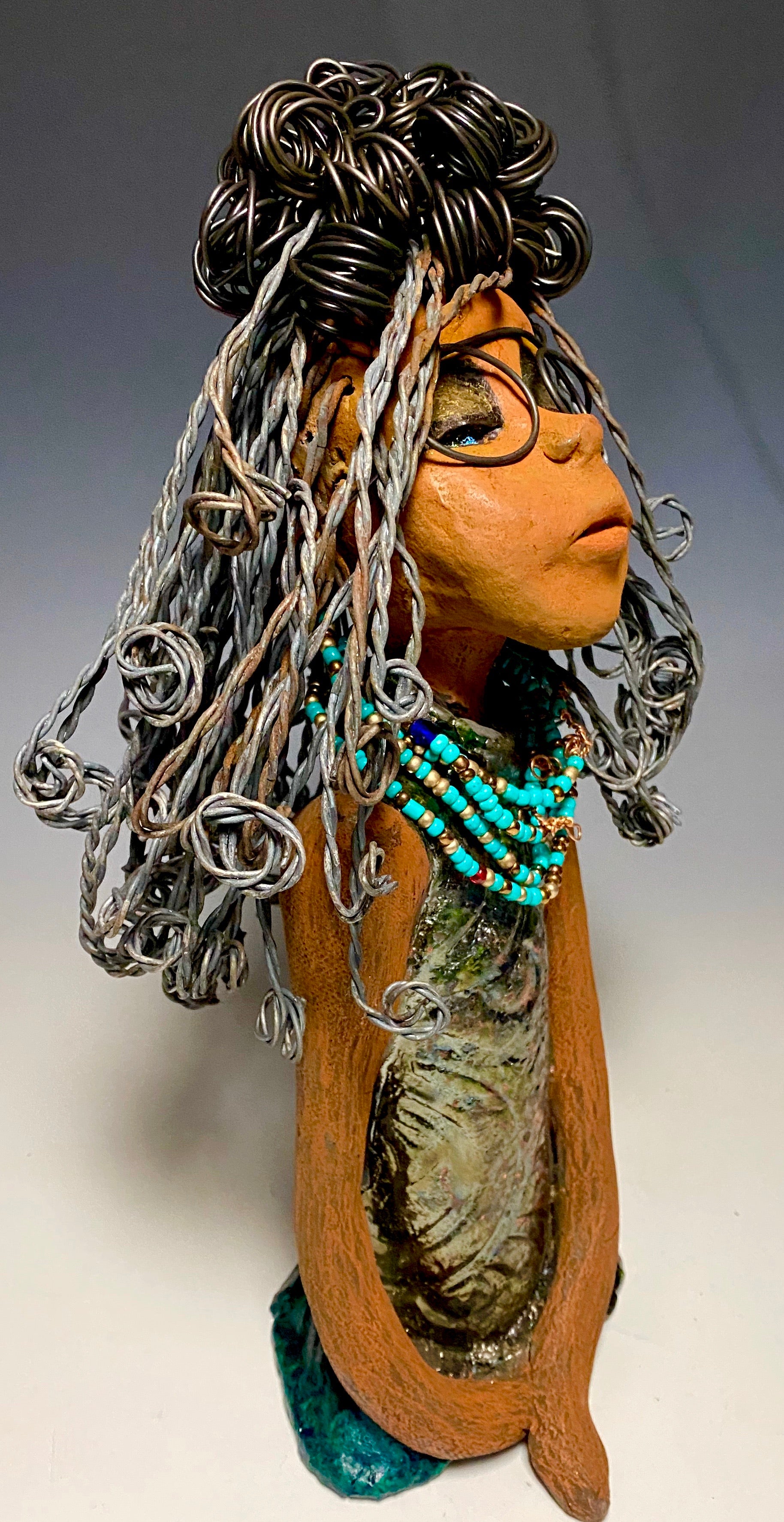 Meet Clarice! Clarice stand 13" x 6" x 5" and weighs 4.03 lbs. She has a lovely honey brown complexion with big bright eyes. Clarice wears glasses now! Her Herdew took over 2 hours to complete. It's made with 16 gauge wire. Clarice's dress is alligator green with streaks of copper. She has a blue beaded spiral necklace around her neck. Clarice looks to have a New ExtrArtindary Vision for 2021!