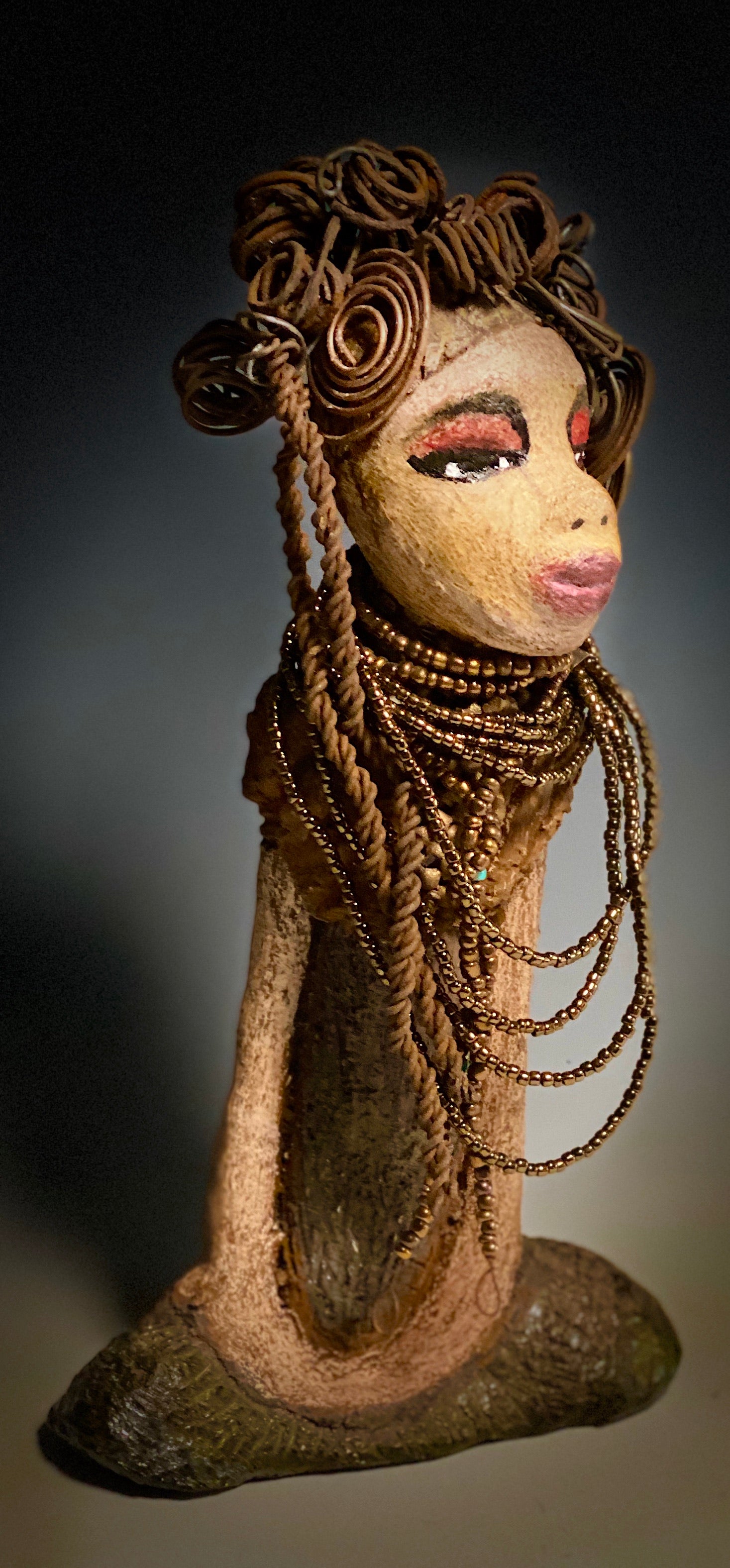You will love having Cardi around! Cardi stands 11" x 5.5" x 3" and weighs 1.14 lbs. She has an awesome honey beige complexion. Cardi wears a dark metallic copper dress with a chocolate ruffle scarf.  She wears a multitude of amber colored hand strung beads. I do believe Cardi would like it if you gave her a space in your home!