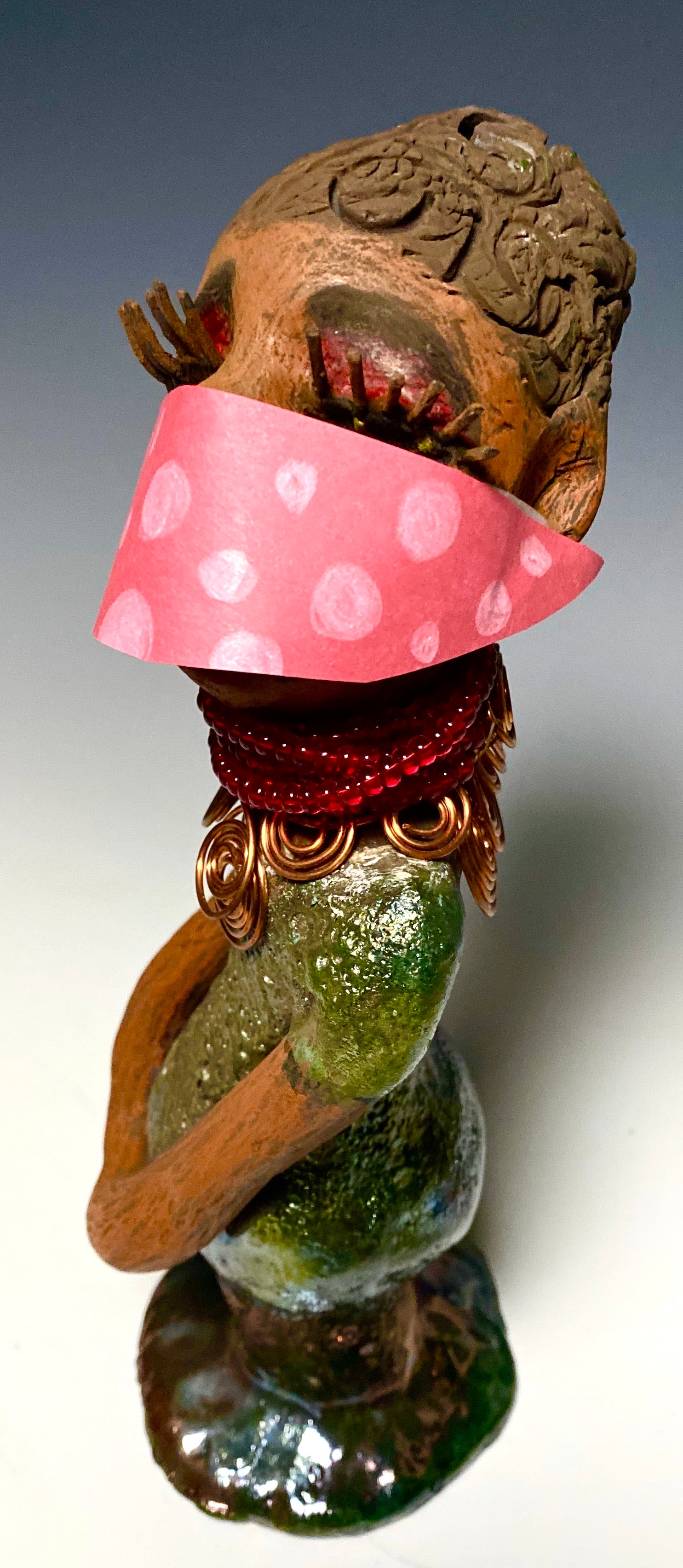 Meet Mona! Mona stands 11.5" x 4" x 4" and weighs 1.81 lbs. She has a lovely chocolate brown complexion with chocolate brown lips. She has a braided hairstyle.  Mona has a colorful metallic antique copper glazed dress. She wears a spiral copper wire necklaces on top of a beaded ruby red collar. With long lashes and eyes wide opened, Mona has hopes of finding a new home. You may have noticed, Mona is the one of a few herdew  sculptures that wears a face mask during this unprecedented time.