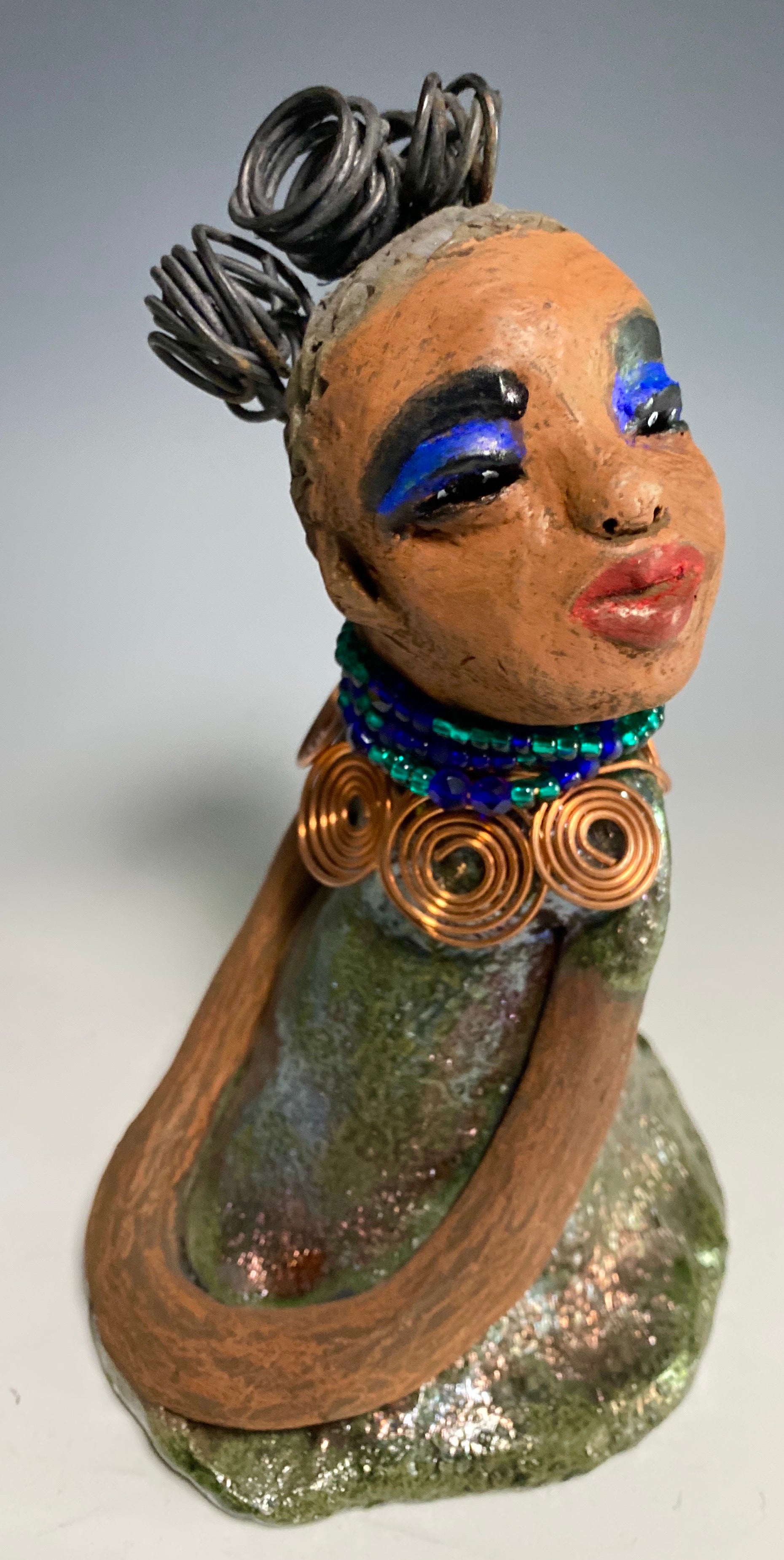 Meet Nia! Nia stands 8" x 6" x 6" and weighs 1.08 lbs. She has a lovely honey brown complexion with  reddish brown lips. She has a braided  wire bun hairstyle.  Nia has a colorful metallic green antique copper glazed dress. She wears a spiral copper wire necklaces with green beads underneath.  With eyes wide opened, Nia has hopes of finding her way into your home.