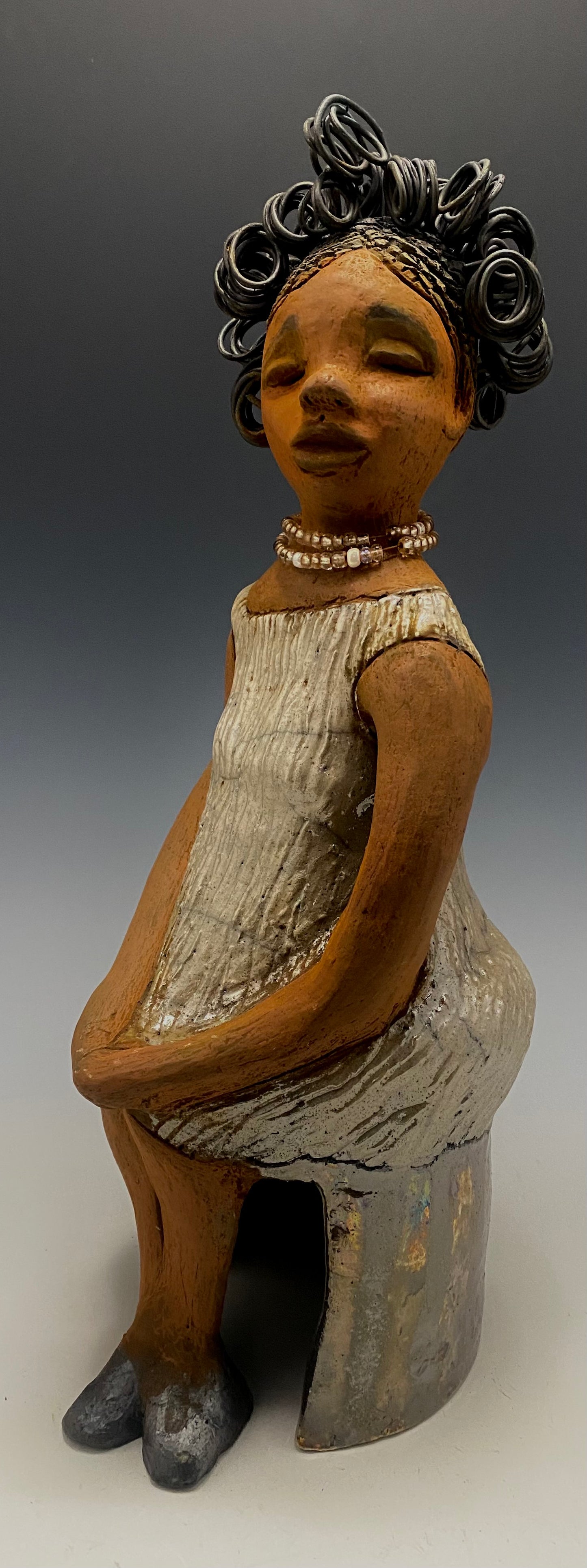 Meet Gwen! You know someone like Gwen. One who reaps elegance and sophistication! Gwen stands 13 " x 6" x 6" and weighs 3.07 lbs. She has a lovely brown complexion with over 5 feet of 16 gauge spiral coiled wire hair. Gwen's metallic off white dress is accented by a multicolored white and amber beaded necklace. Gwen’s straight face gives her a distinguished no nonsense look. Gwen sits with her hands rested as she awaits a special place to grace your home.
