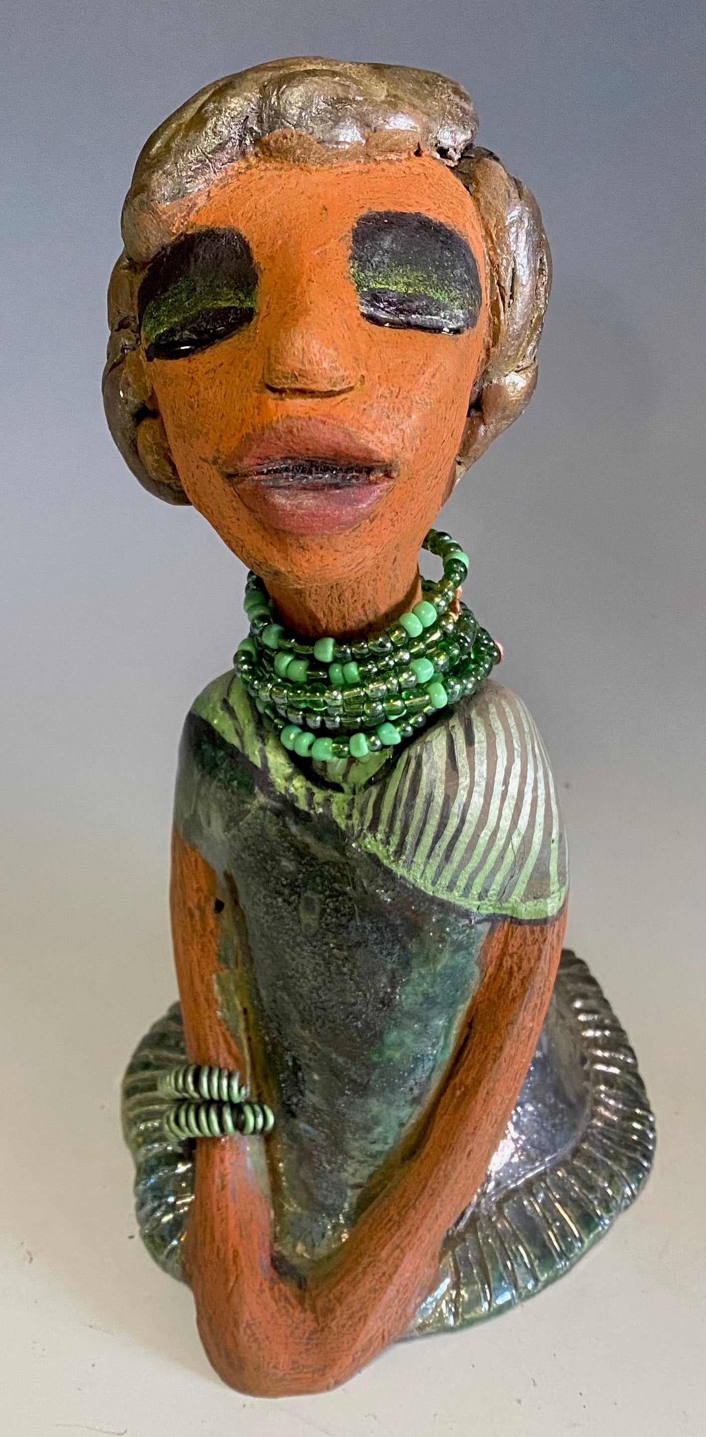 Meet Cora! Cora stands 9" x 5" x 6" and weighs 1.12 lbs. She has a lovely beige brown complexion with long etched braided clay hair. Cora’s long loving arms rest beside her copper green dress. Her dress is alligator green with a green coiled arm band. Cora wears a long string mixed green beaded necklace. Cora is a sophisticated lady that will grace your home.