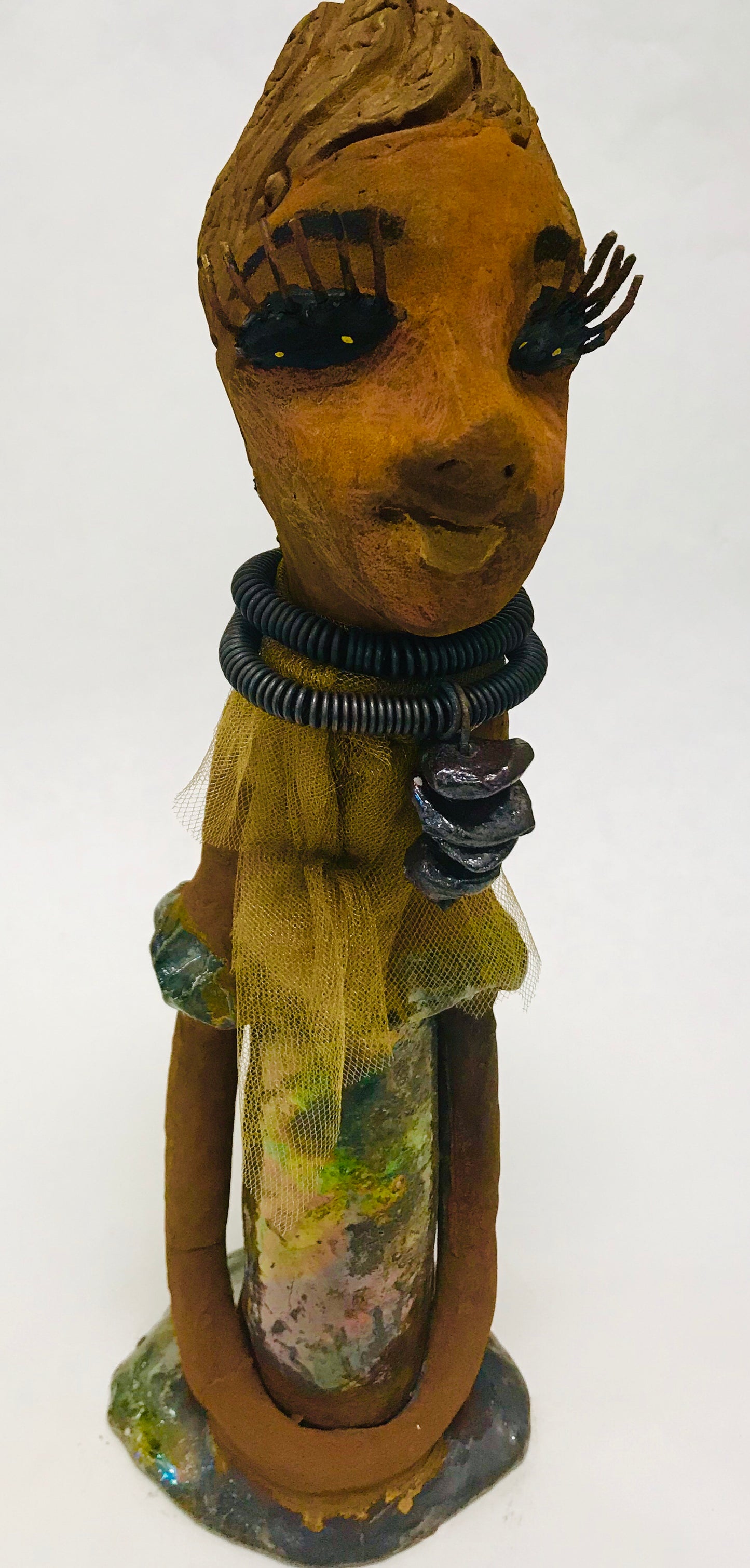 Trinity stands 14" x 4" x 4" and weighs 2.7 lbs. She has a lovely honey brown complexion with reddish brown lips. She has a braided hairstyle. Trinity has a colorful metallic green antique copper glazed dress. She wears a spiral wire necklaces with raku beads underneath an antique gold tulle.. With long lashes and eyes wide opened, Trinity has hopes of finding a new home.