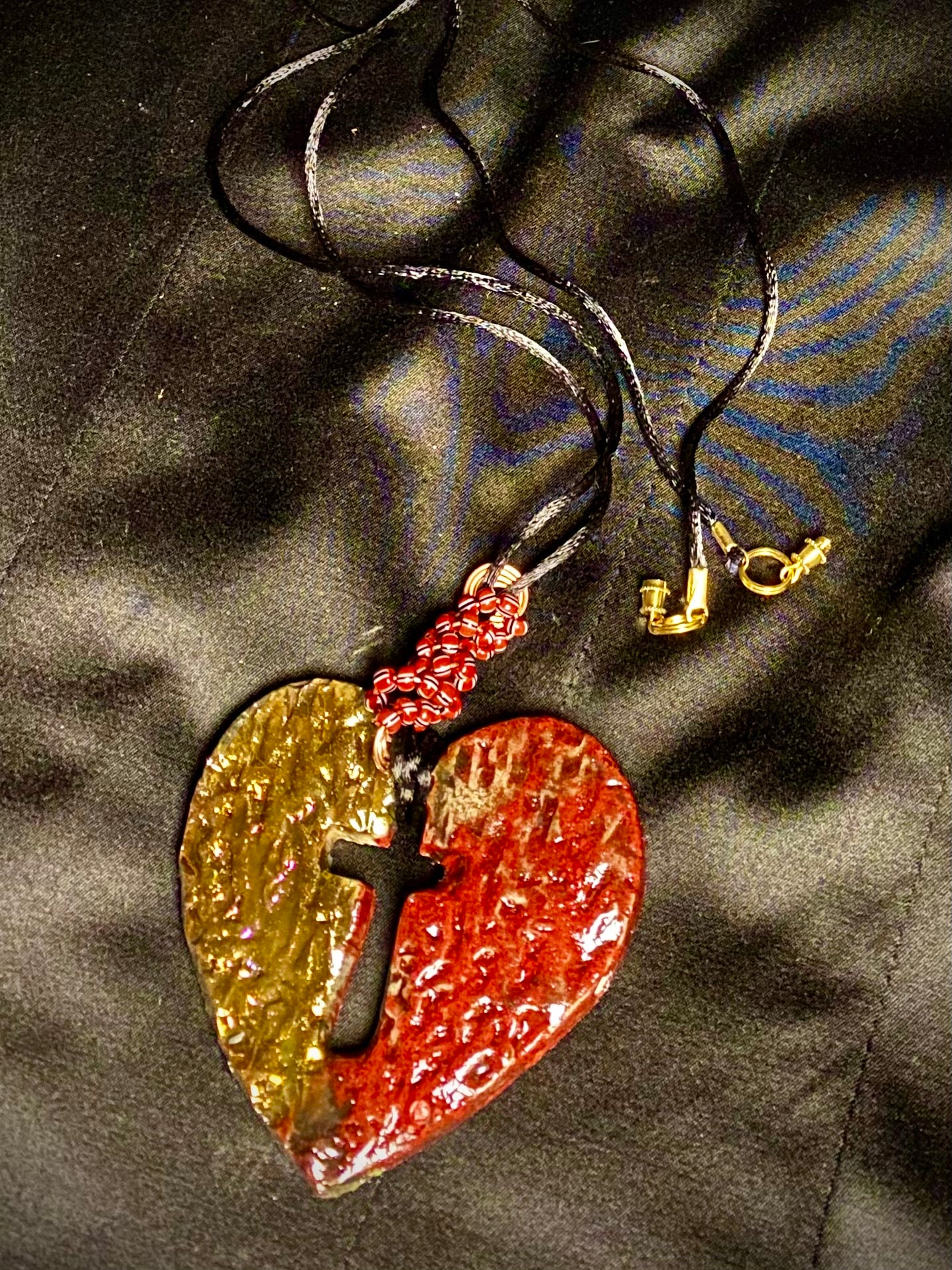 Have A Heart ! Each heart pendant is handmade with love! It is 3"x 3" and weighs approx. 3ozs. This pendant has a red and gold metallic raku glazes that renders a unique translucent patina. The heart is textured and has a cut out cross in the center. It holds a spiral of red and white mini beads on a spiral copper wire. This pendant has a nice 12" black suede cord!