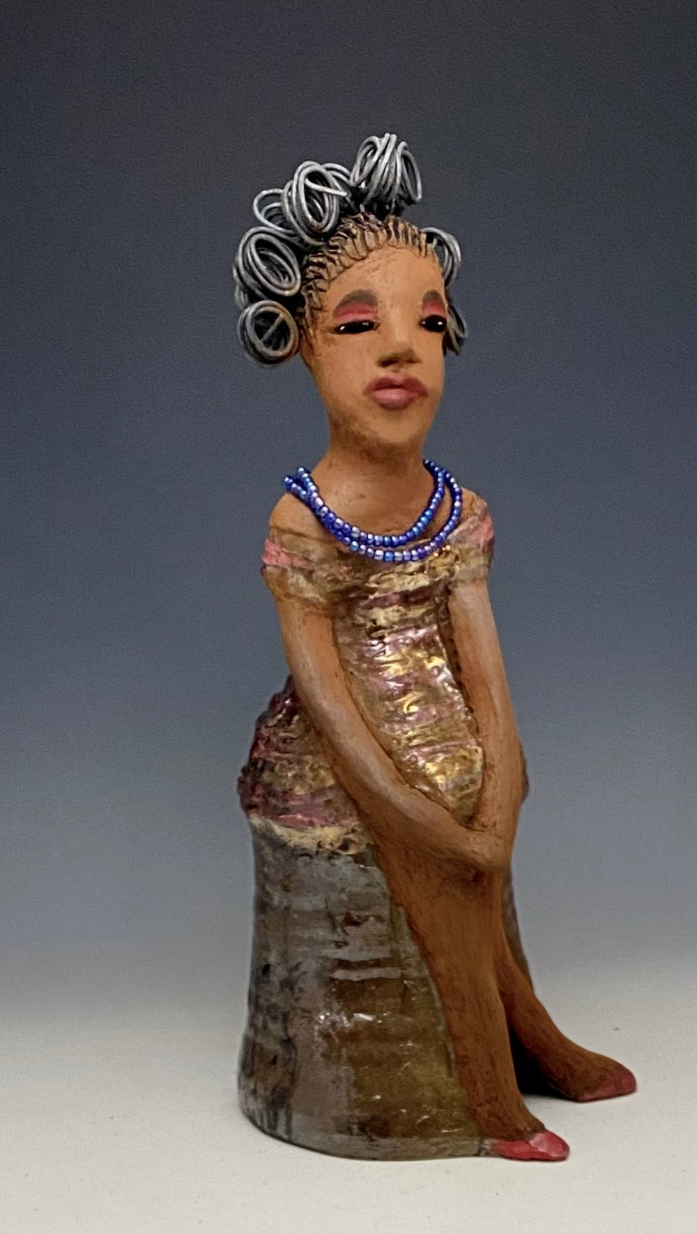 You know someone like Kim. One who reaps elegance and sophistication! Kim stands 13" x 5" x 7" and weighs 2.07 lbs. She has a lovely light beige complexion with over 4 feet of 16 gauge spiral coiled wire hair. Kim's metallic copper red and gold dress is accented by a multicolored aqua blue beaded necklace. Kim's maroon eye shadow gives her a distinguished mystical look. One must wonder what is Kim thinking? Kim sits with her legs crossed as she awaits a special place to grace your home.