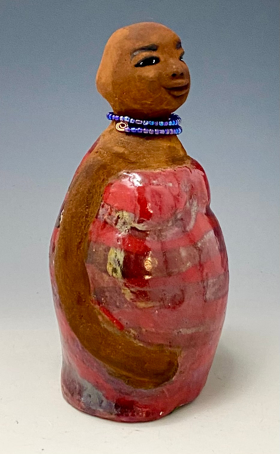 Meet Cora! Cora stands 9" x 7" x 4.5" and weighs  2lbs. She has a lovely honey brown complexion with lovely reddish brown lips. Cora wears a clay bald hairstyle. Cora has a striped heart red and gold glazed dress. She wears a blue beaded necklace. With eyes wide opened, Janae' has hopes of finding a new home.