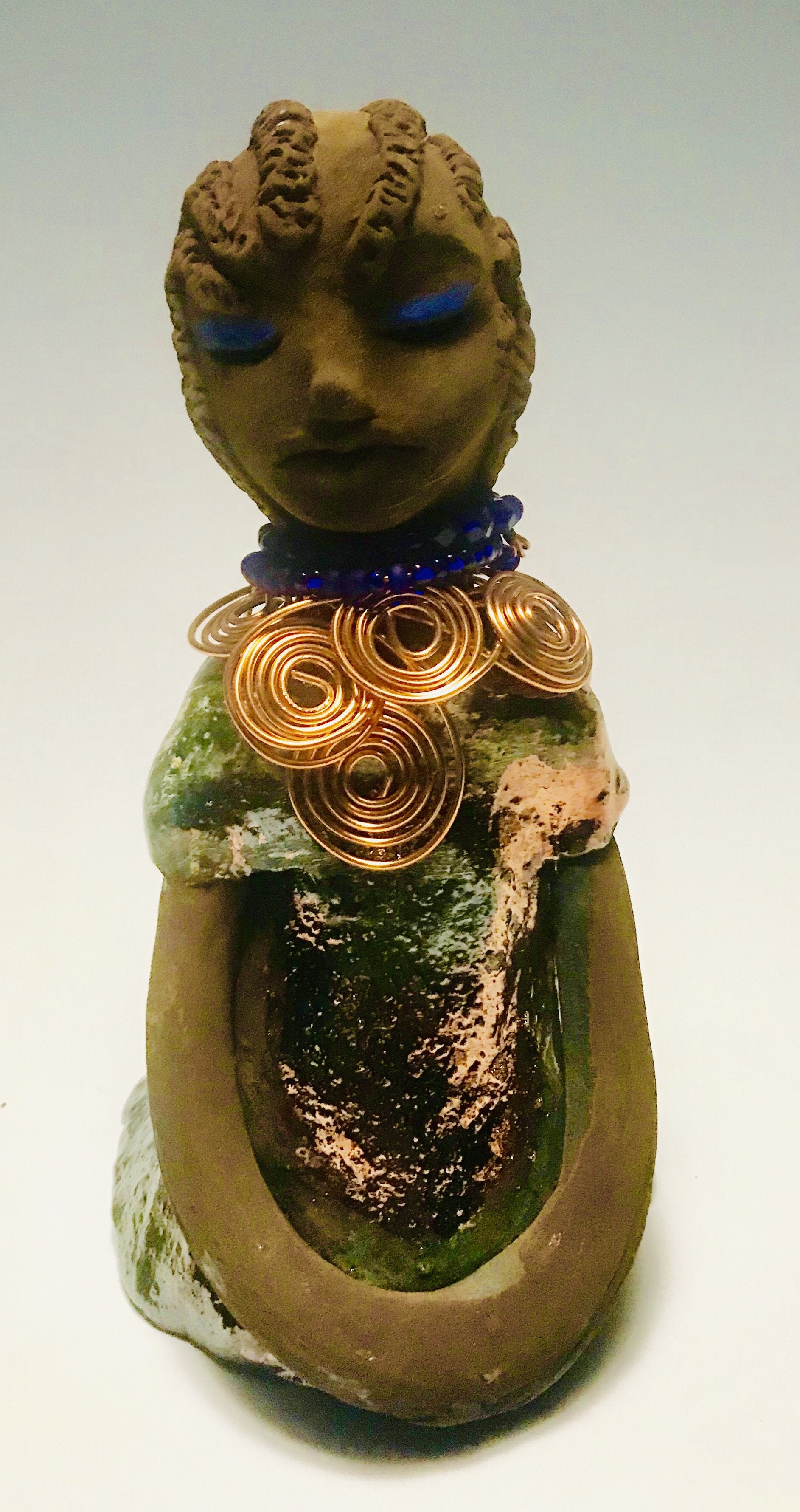 Anja stands 7" x 4" x 4" and weighs 1.01 lbs. She has a lovely honey brown complexion with  reddish brown lips. She has a braided hairstyle.  Anja has a colorful metallic green antique copper glazed dress. She wears a spiral copper wire necklace on top of a blue beaded collar. With eyes slightly opened, Anja has hopes of finding a new home.