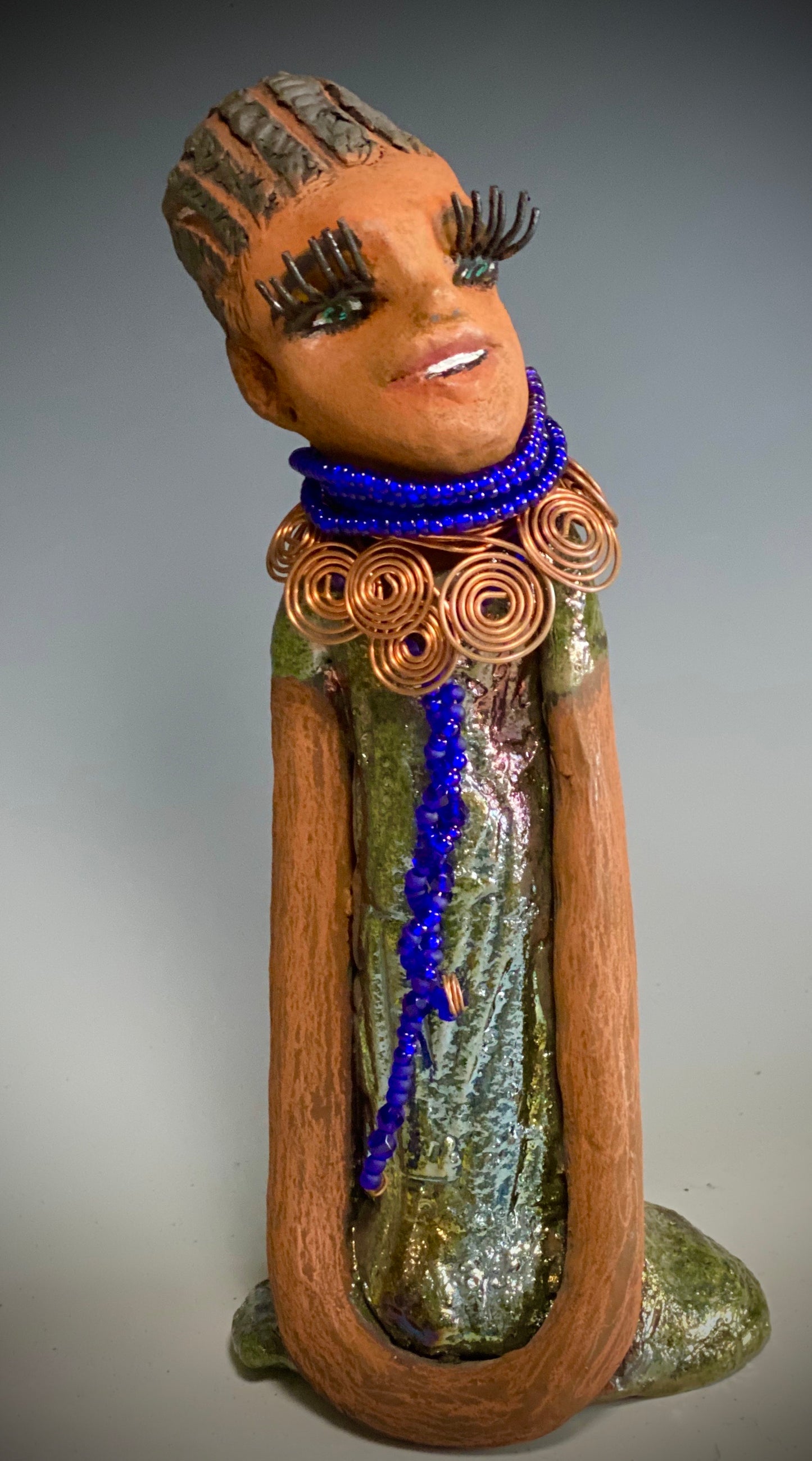 Meet Ophelia! Ophelia stands 7.5" x 4" x 5" and weighs 1.02 lbs. She has a lovely honey brown complexion with reddish brown lips. She has a short braided hairstyle. Ophelia has a colorful metallic antique copper glazed dress. She wears spiral copper wire necklaces on top of an aqua blue beaded collar. Ophelia long loving arms rest at her side. With accent blue eye shadow and eyes wide opened, Ophelia has hopes of finding her way into your home!