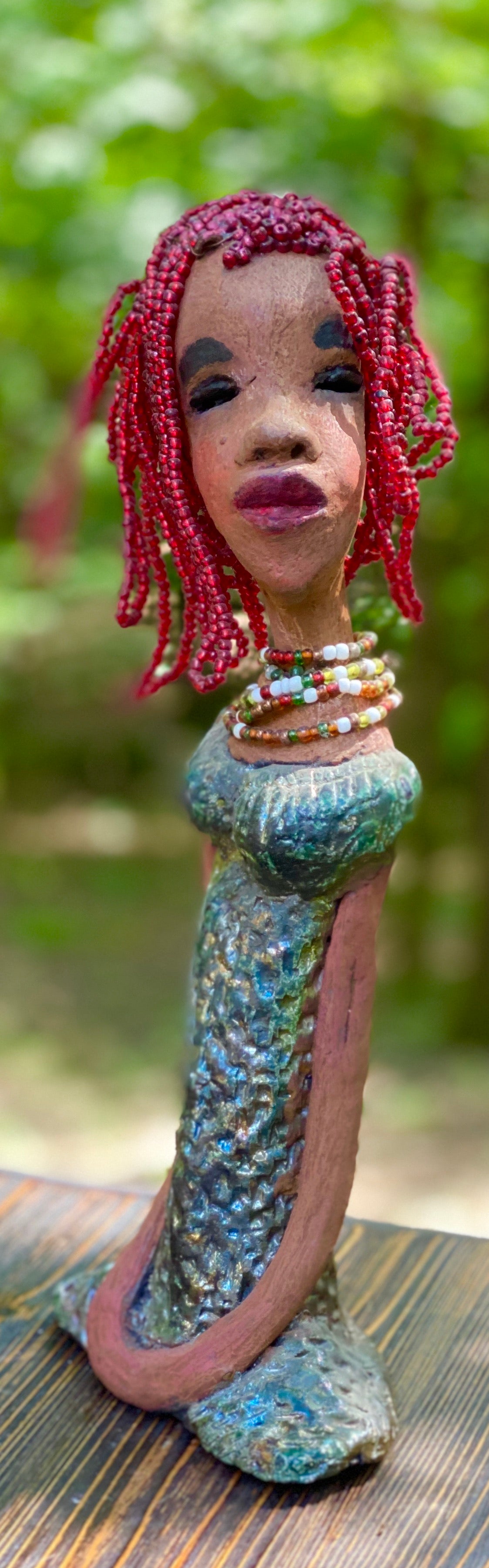 Meet July! July stands 14" x 5" x 3" and weighs 2.05 lbs. July has a head full of red beaded hair. She has a honey brown complexion. July dress has a metallic textured copper green. She wears a multi colored beaded necklace. With her head slightly turned her long loving arms gently rest at her side.