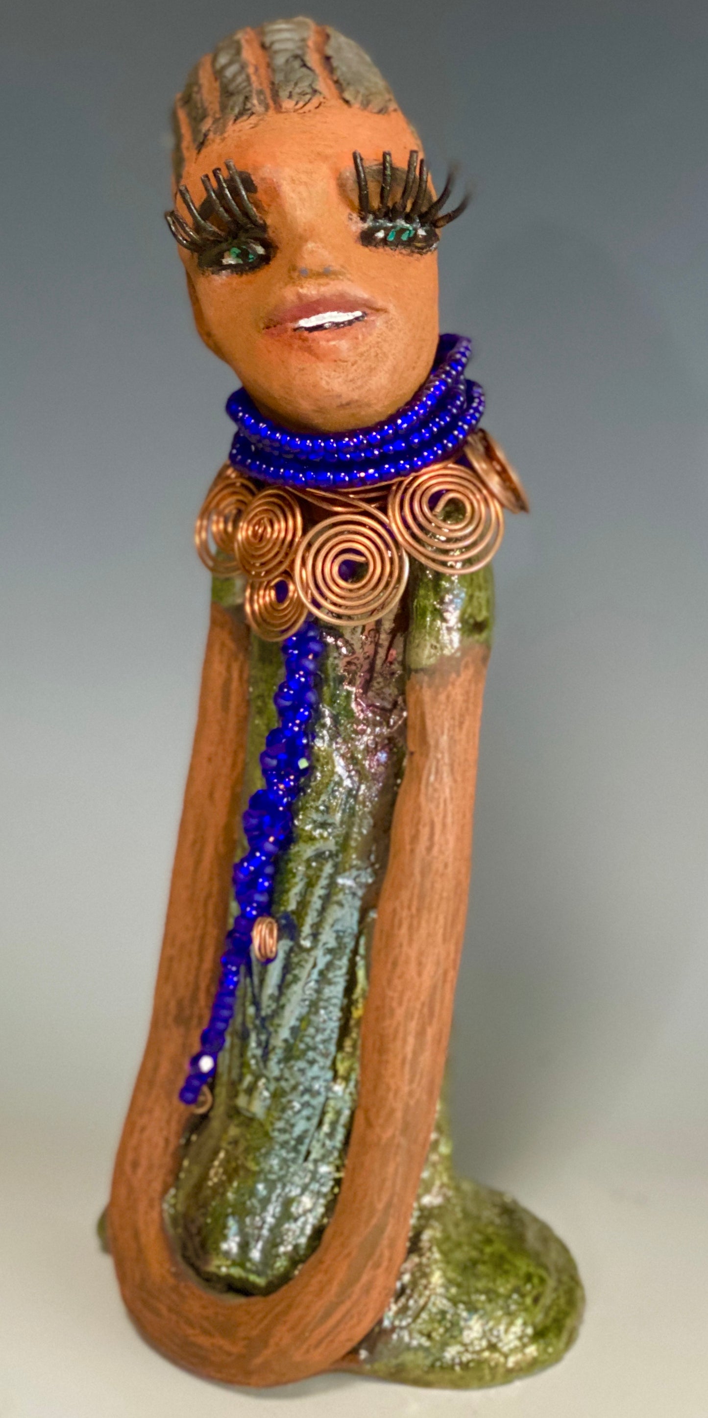 Meet Ophelia! Ophelia stands 7.5" x 4" x 5" and weighs 1.02 lbs. She has a lovely honey brown complexion with reddish brown lips. She has a short braided hairstyle. Ophelia has a colorful metallic antique copper glazed dress. She wears spiral copper wire necklaces on top of an aqua blue beaded collar. Ophelia long loving arms rest at her side. With accent blue eye shadow and eyes wide opened, Ophelia has hopes of finding her way into your home!