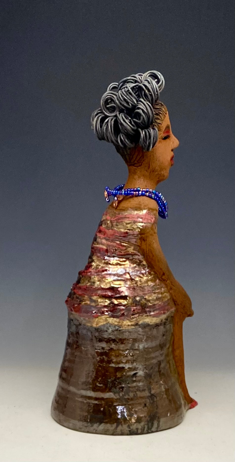 You know someone like Kim. One who reaps elegance and sophistication! Kim stands 13" x 5" x 7" and weighs 2.07 lbs. She has a lovely light beige complexion with over 4 feet of 16 gauge spiral coiled wire hair. Kim's metallic copper red and gold dress is accented by a multicolored aqua blue beaded necklace. Kim's maroon eye shadow gives her a distinguished mystical look. One must wonder what is Kim thinking? Kim sits with her legs crossed as she awaits a special place to grace your home.