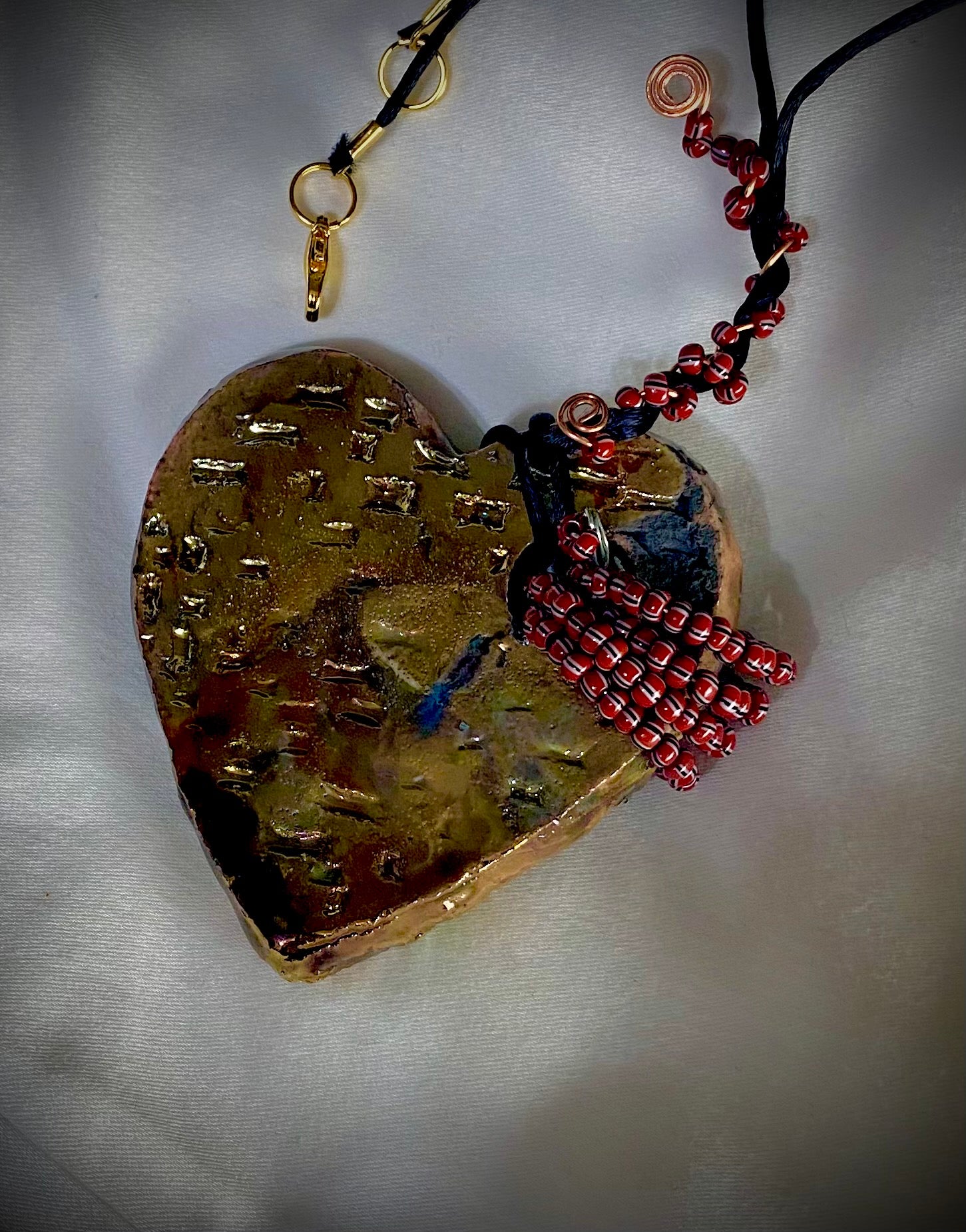 Have A Heart ! Each heart pendant is handmade with love! It is 3"x 3" and weighs approx. 3ozs. This pendant has a white and gold metallic raku glazes that renders a unique translucent  patina. The heart has a textured with cut out cross pattern . It holds a spiral of multicolored mini beads on a spiral copper wire. This pendant has a nice 12" black suede cord!