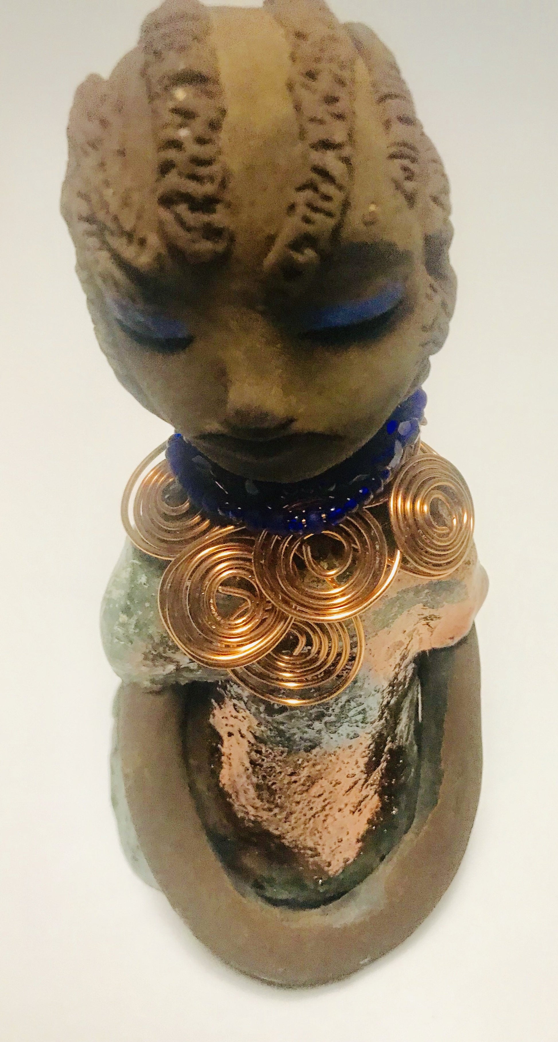 Anja stands 7" x 4" x 4" and weighs 1.01 lbs. She has a lovely honey brown complexion with  reddish brown lips. She has a braided hairstyle.  Anja has a colorful metallic green antique copper glazed dress. She wears a spiral copper wire necklace on top of a blue beaded collar. With eyes slightly opened, Anja has hopes of finding a new home.