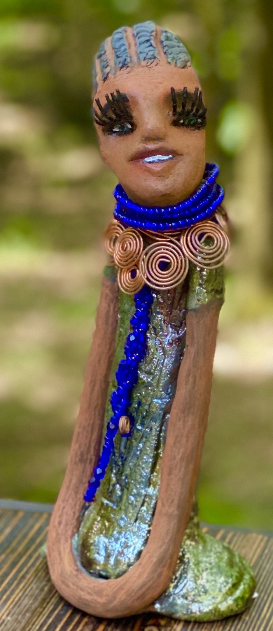Meet Ophelia! Ophelia stands 7.5" x 4" x 5" and weighs 1.02 lbs. She has a lovely honey brown complexion with reddish brown lips. She has a short braided hairstyle. Ophelia has a colorful metallic antique copper glazed dress. She wears spiral copper wire necklaces on top of an aqua blue beaded collar. Ophelia long loving arms rest at her side. With accent blue eye shadow and eyes wide opened, Ophelia has hopes of finding her way into your home!