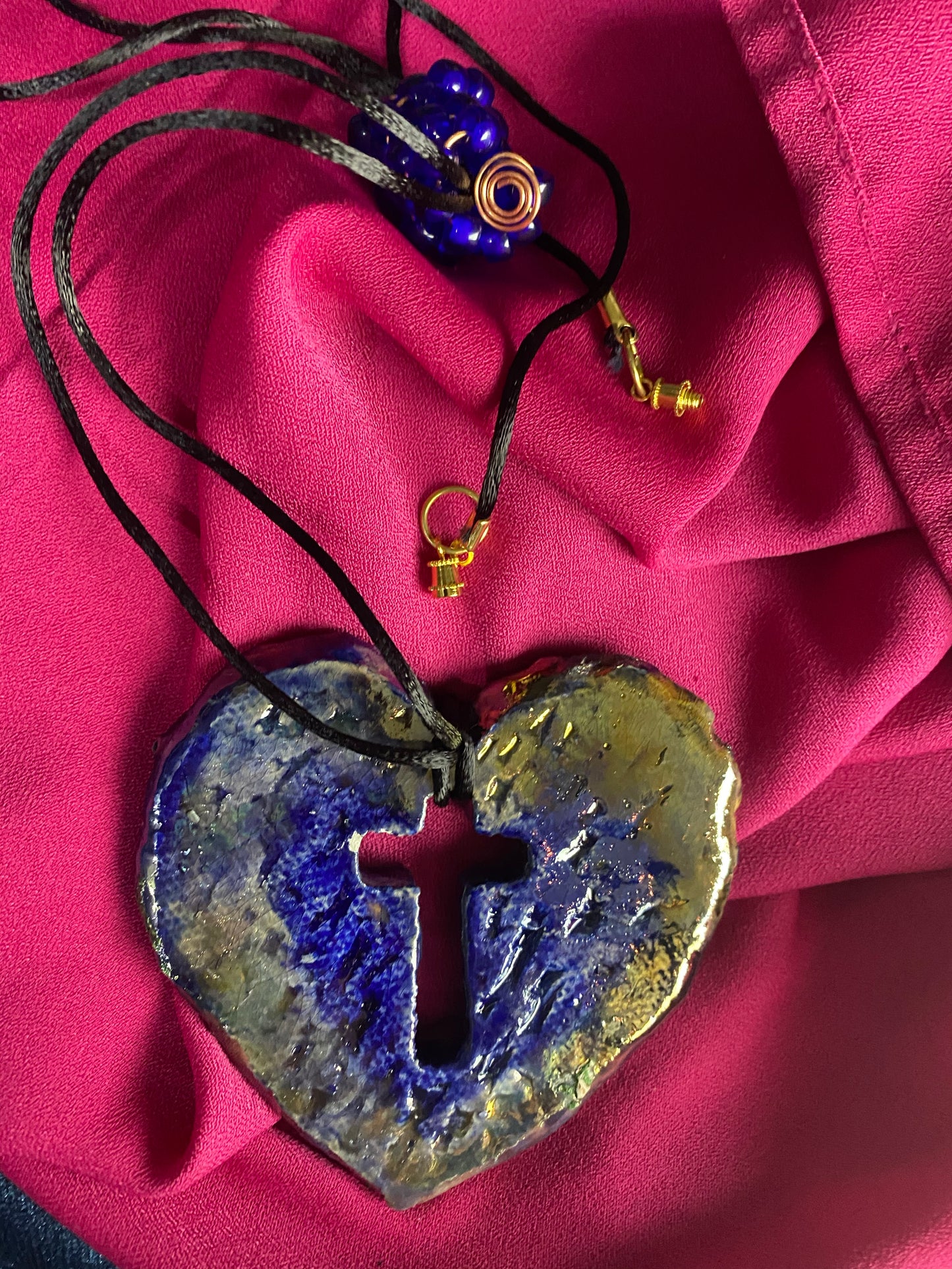 Have A Heart ! Each heart pendant is handmade with love! It is 3"x 3" and weighs approx. 3ozs. This pendant has a royal blue and gold metallic raku glazes that renders a unique translucent patina. The heart has a cut out cross in the center with a textured pattern. It holds a spiral of royal blue mini beads on a spiral copper wire. This pendant has a nice 12" black suede cord!
