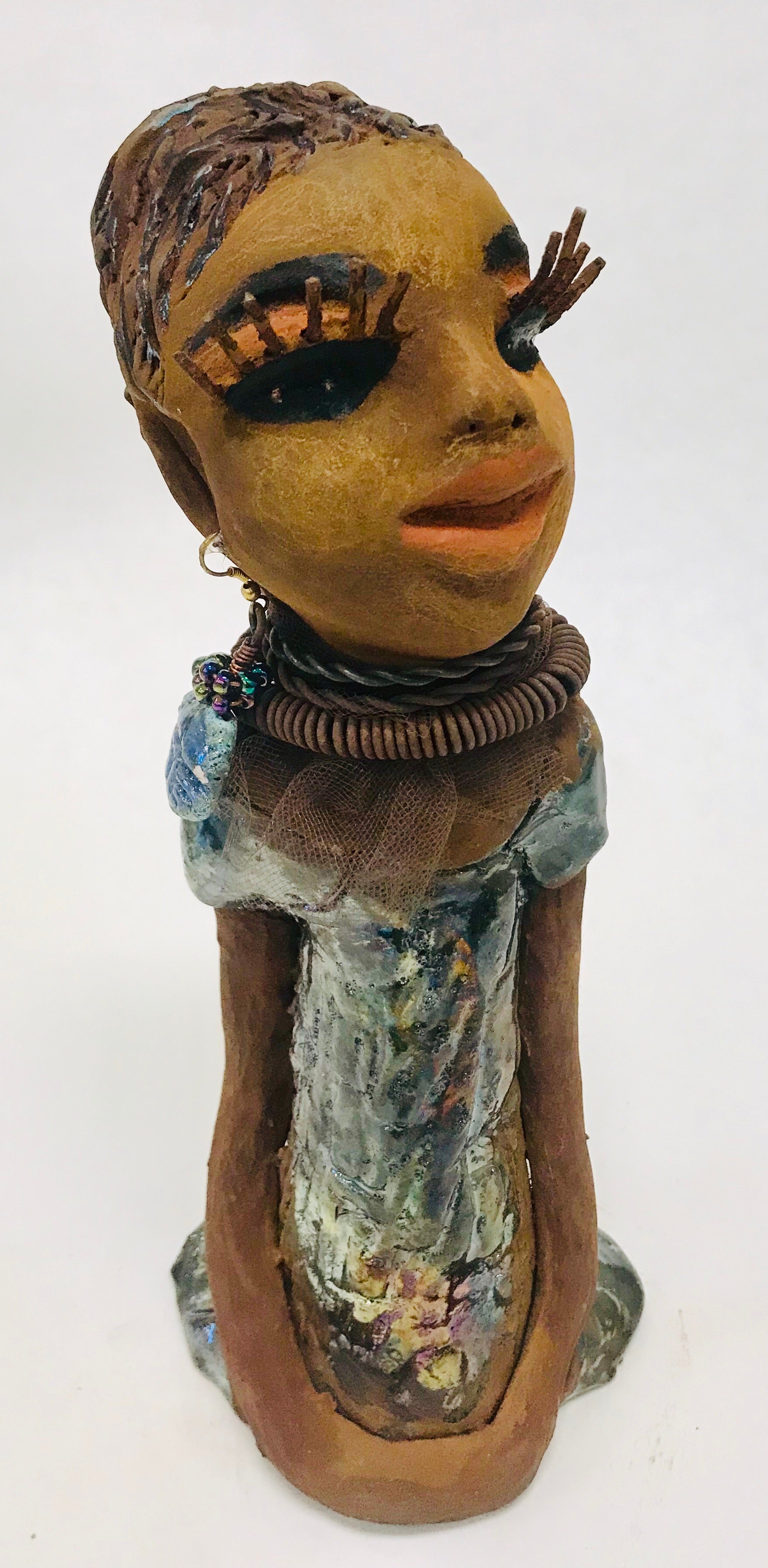 Sorbari stands 11" x 4" x 4" and weighs 2.5 lbs. She has a lovely honey brown complexion with  reddish brown lips. She has a braided hairstyle.  Sorbari has a colorful metallic green antique copper glazed dress. She wears two spiral wire necklaces. With long eyes slightly opened, Sorbari has hopes of finding a new home.