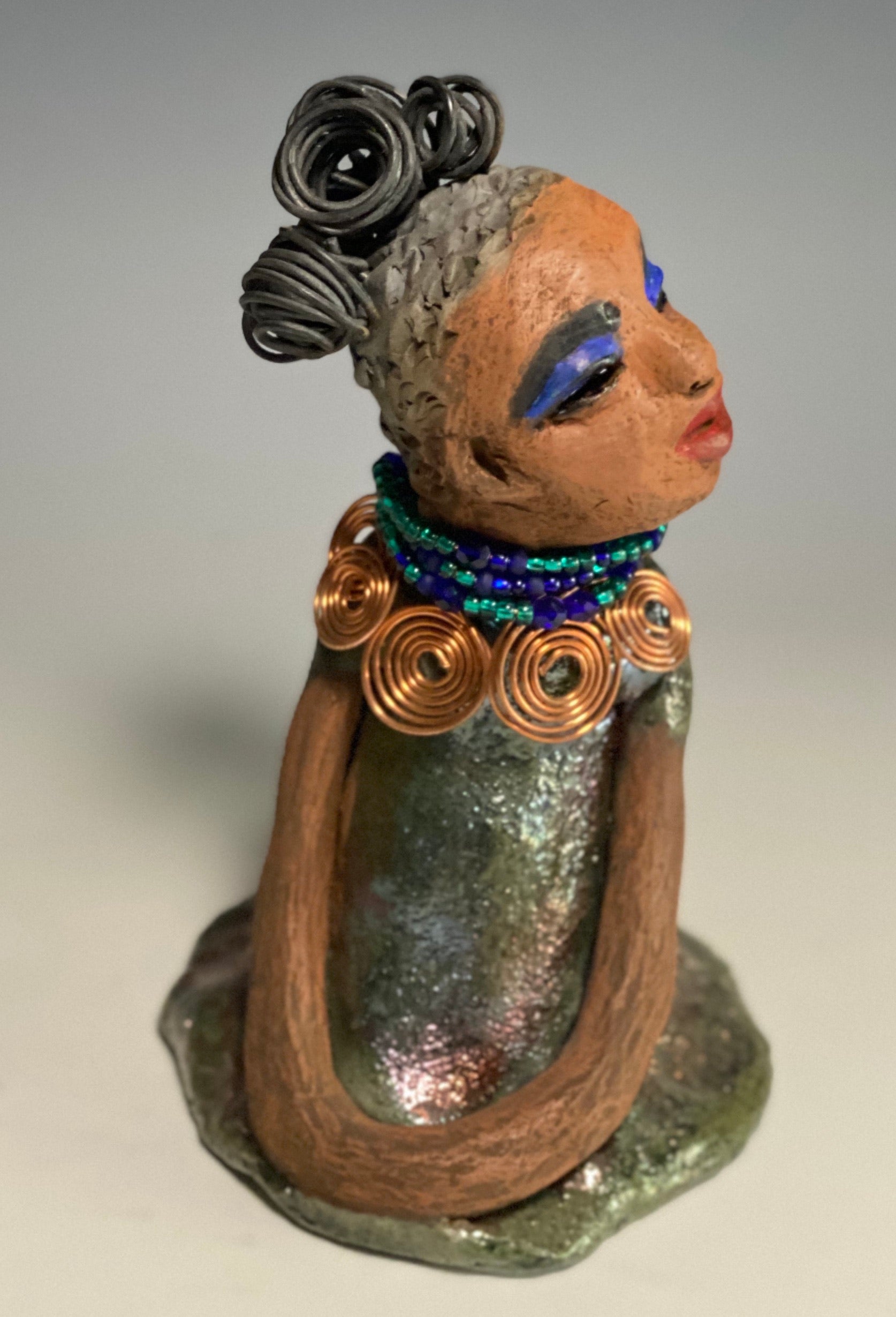 Meet Nia! Nia stands 8" x 6" x 6" and weighs 1.08 lbs. She has a lovely honey brown complexion with  reddish brown lips. She has a braided  wire bun hairstyle.  Nia has a colorful metallic green antique copper glazed dress. She wears a spiral copper wire necklaces with green beads underneath.  With eyes wide opened, Nia has hopes of finding her way into your home.