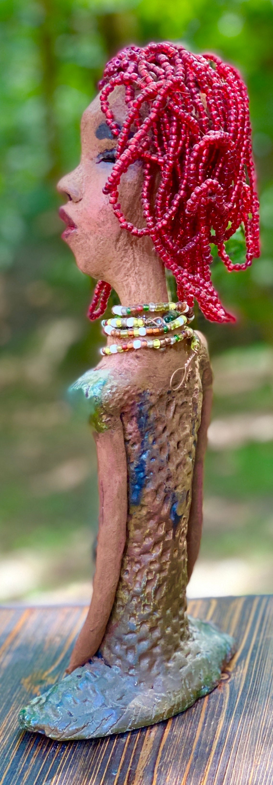 Meet July! July stands 14" x 5" x 3" and weighs 2.05 lbs. July has a head full of red beaded hair. She has a honey brown complexion. July dress has a metallic textured copper green.  She wears a multi colored beaded necklace. With her head slightly turned her long loving arms gently rest at her side.