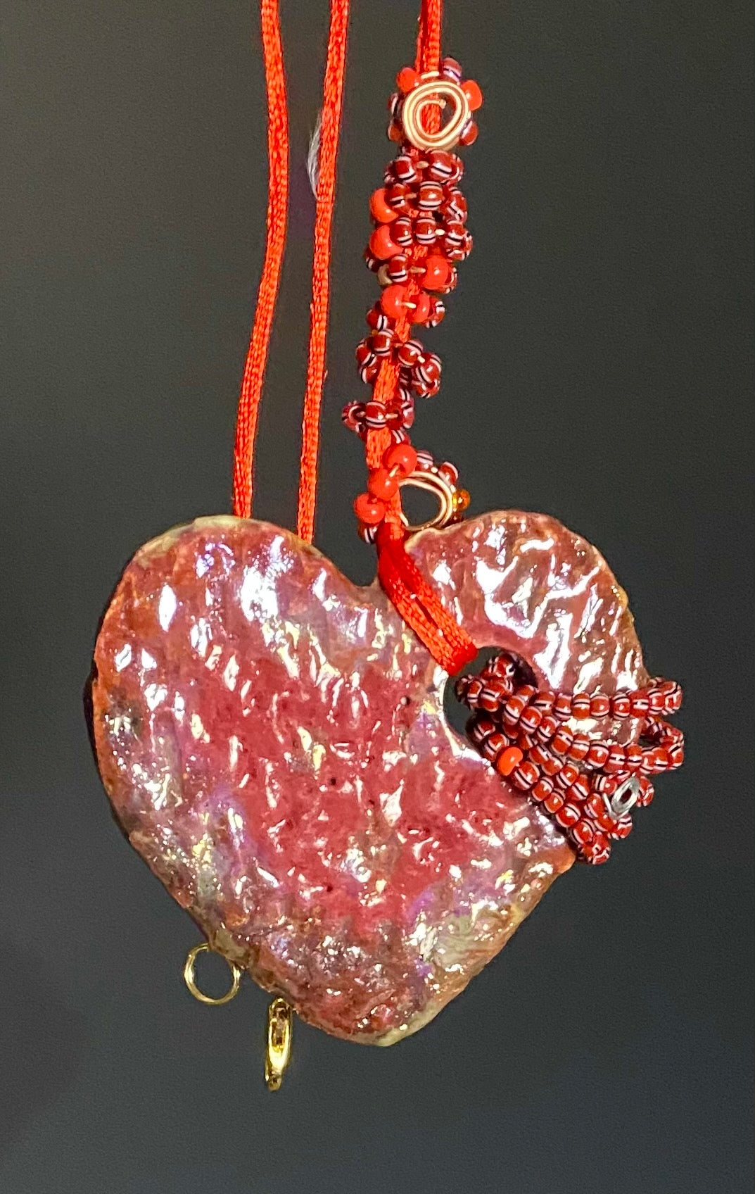 Have A Heart! Each heart pendant is handmade with love! It is 3"x 3"and weighs approx. 3ozs. This pendant has a red, white, and gold metallic raku glazes that renders a unique translucent  patina. The heart has a textured pattern . Both sides are  are different and equally beautiful! It holds a spiral of red  and deep purple mini beads on a spiral copper wire. This pendant has a nice 12" red adjustable rattail cord!
