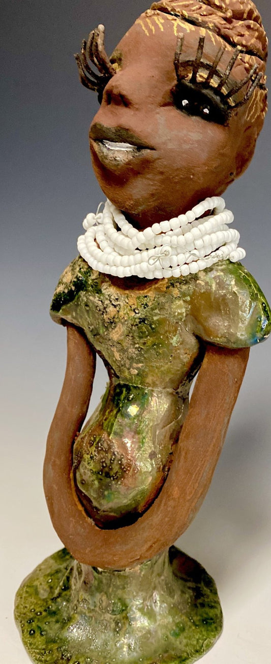 Meet Erma! Erma stands 11.5" x 5" x 5" and weighs 2 lbs. She has a lovely dark brown complexion with brown lips. She has a braided hairstyle.  Erma has a colorful metallic green antique copper glazed dress. She wears a bright white beaded necklace. With long lashes and eyes wide opened and ultra white teeth, Erma has hopes of finding her way into your home!