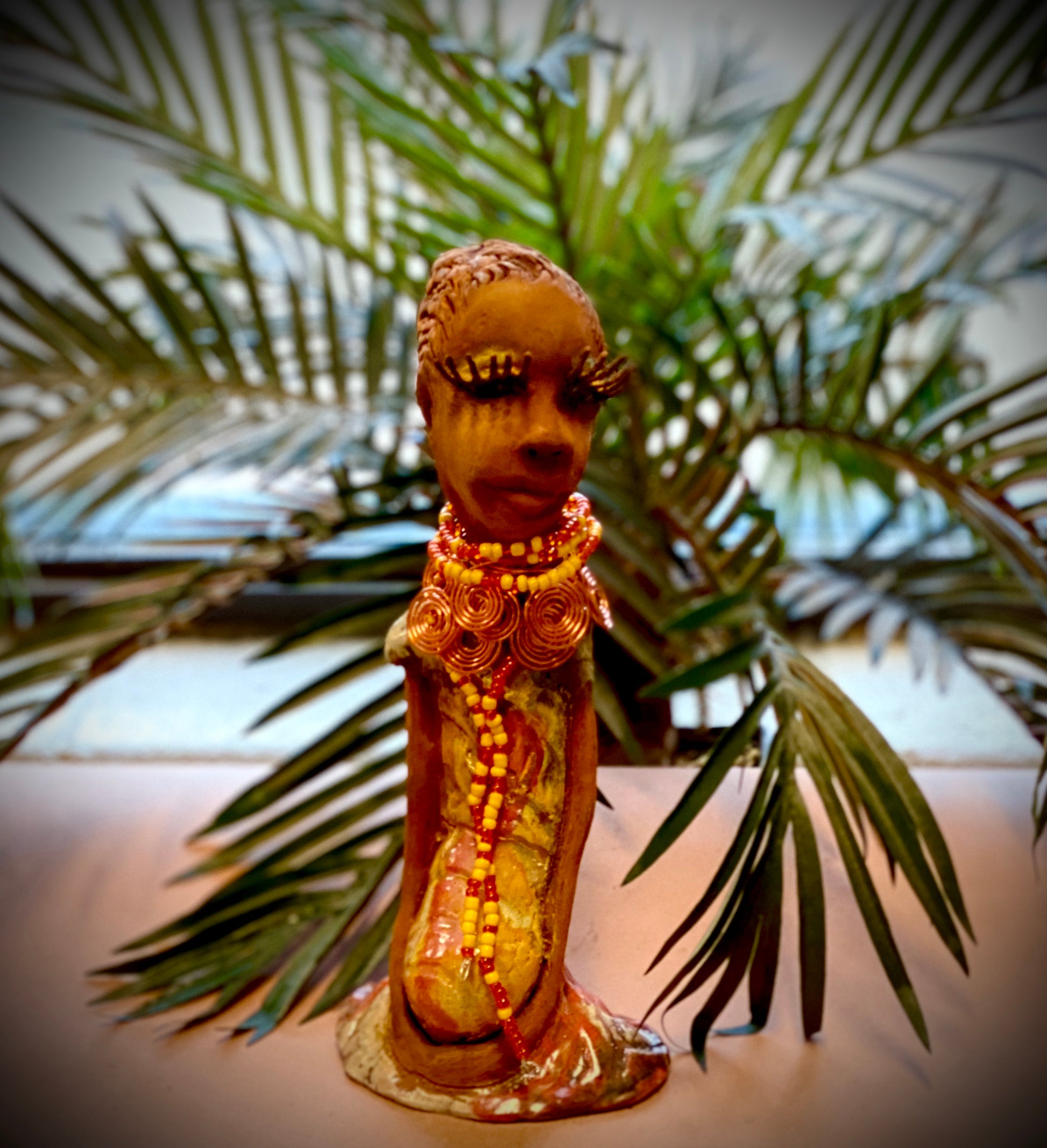 Reandra has a colorful reddish metallic green antique copper glazed dress. She wears a spiral copper wire necklace on top of a yellow red beaded collar. With eyes slightly opened, Reandra has hopes of finding a new home.