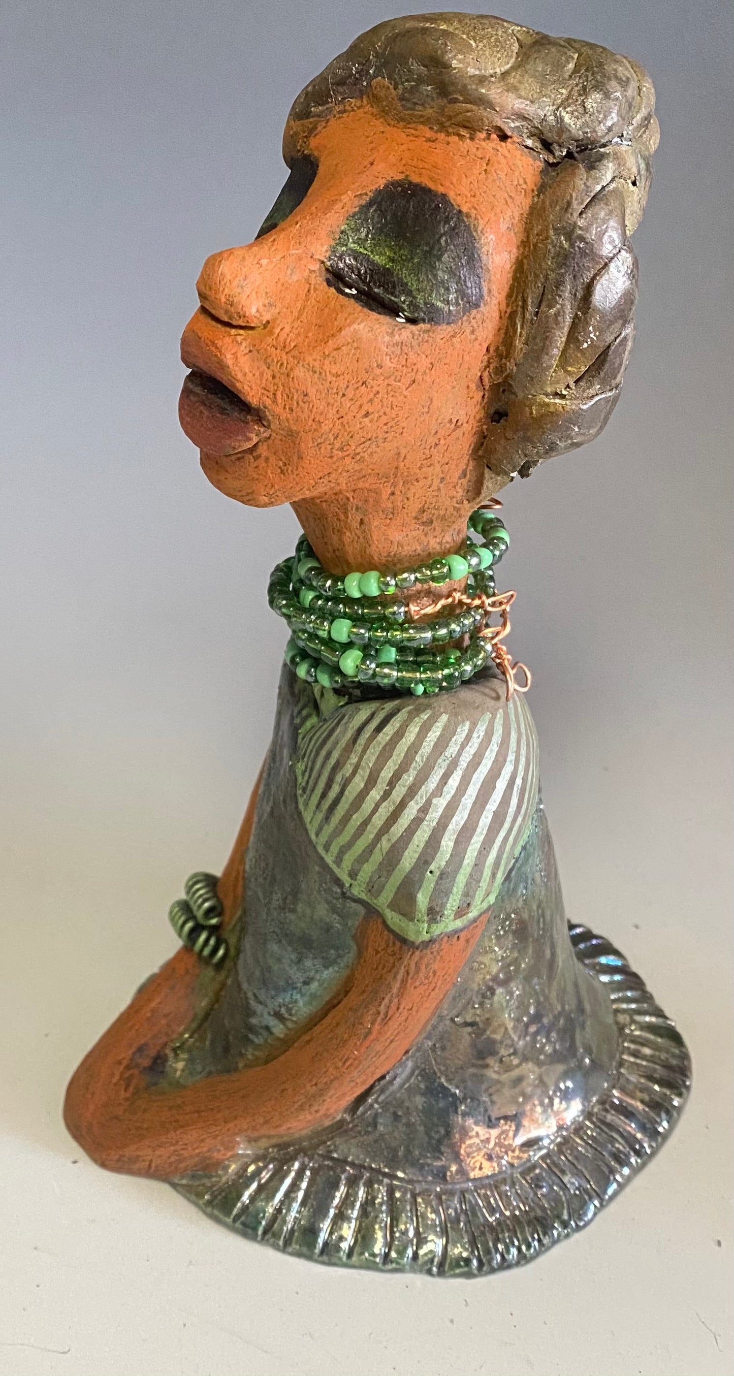 Meet Cora! Cora stands 9" x 5" x 6" and weighs 1.12 lbs. She has a lovely beige brown complexion with long etched braided clay hair. Cora’s long loving arms rest beside her copper green dress. Her dress is alligator green with a green coiled arm band. Cora wears a long string mixed green beaded necklace. Cora is a sophisticated lady that will grace your home.