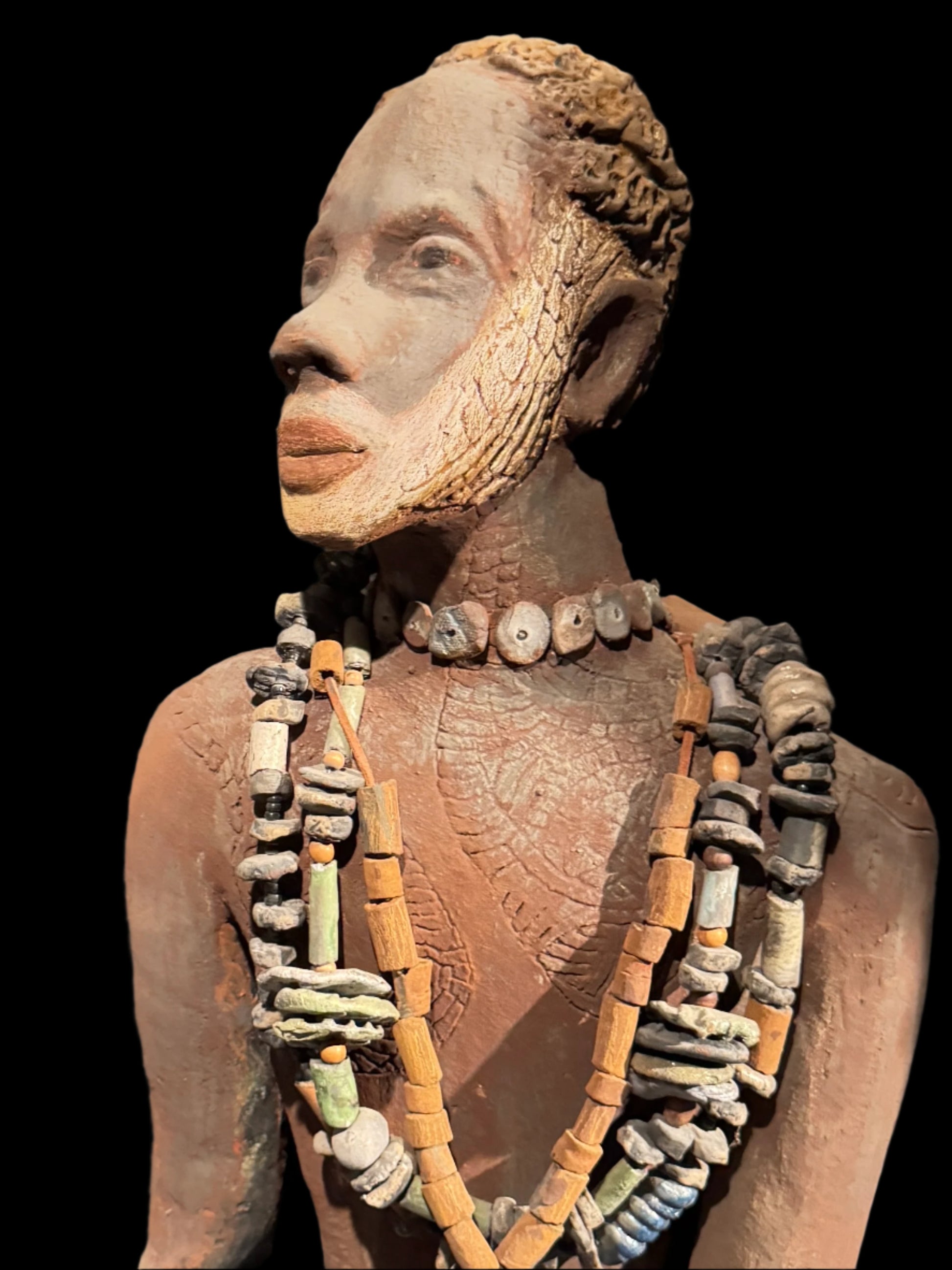  This work of art, created by Lester Jones, is a carefully designed sculpture with rich cultural elements. The piece measures approximately 20" in height and weighs 9 pounds.
The face and chest feature intricate scarification patterns, possibly influenced by African cultural practices. A striking beard and stylized hair suggest symbolic or ceremonial importance.
Furthermore, the figure wears multiple necklaces made from various materials such as clay, stone, and possibly bone or shell. These could signify s