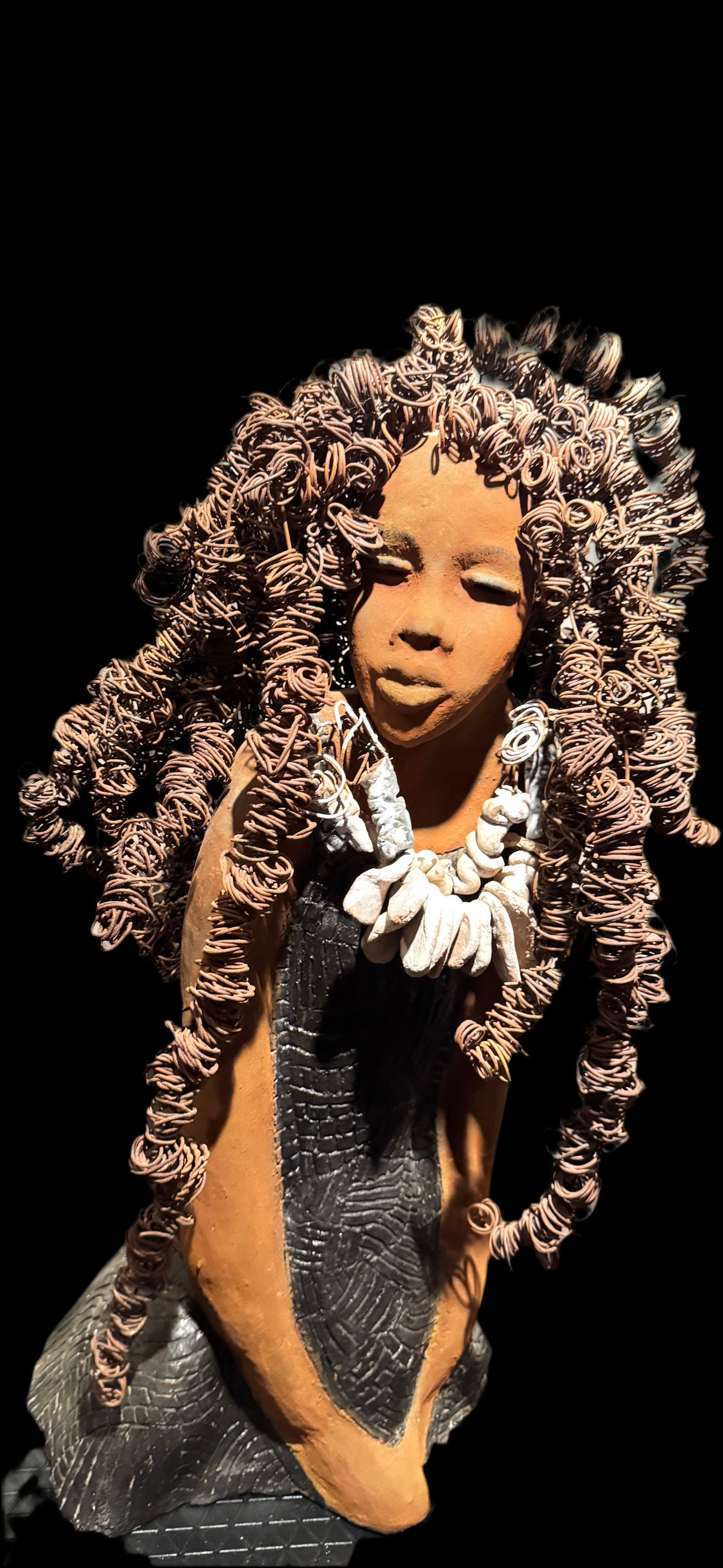 This evocative sculpture by Lester Jones celebrates the beauty, strength, and cultural heritage of African-descended women through form, texture, and symbolism. The figure’s elaborate crown of coiled wire hair evokes vitality, creativity, and a sense of reverence. Her closed eyes and serene expression suggest introspection, grace, or spiritual grounding. Adorned with a necklace  clay shell-like forms and dressed in a patterned robe, the sculpture bridges ancestral tradition and modern artistry.

 

Lester J