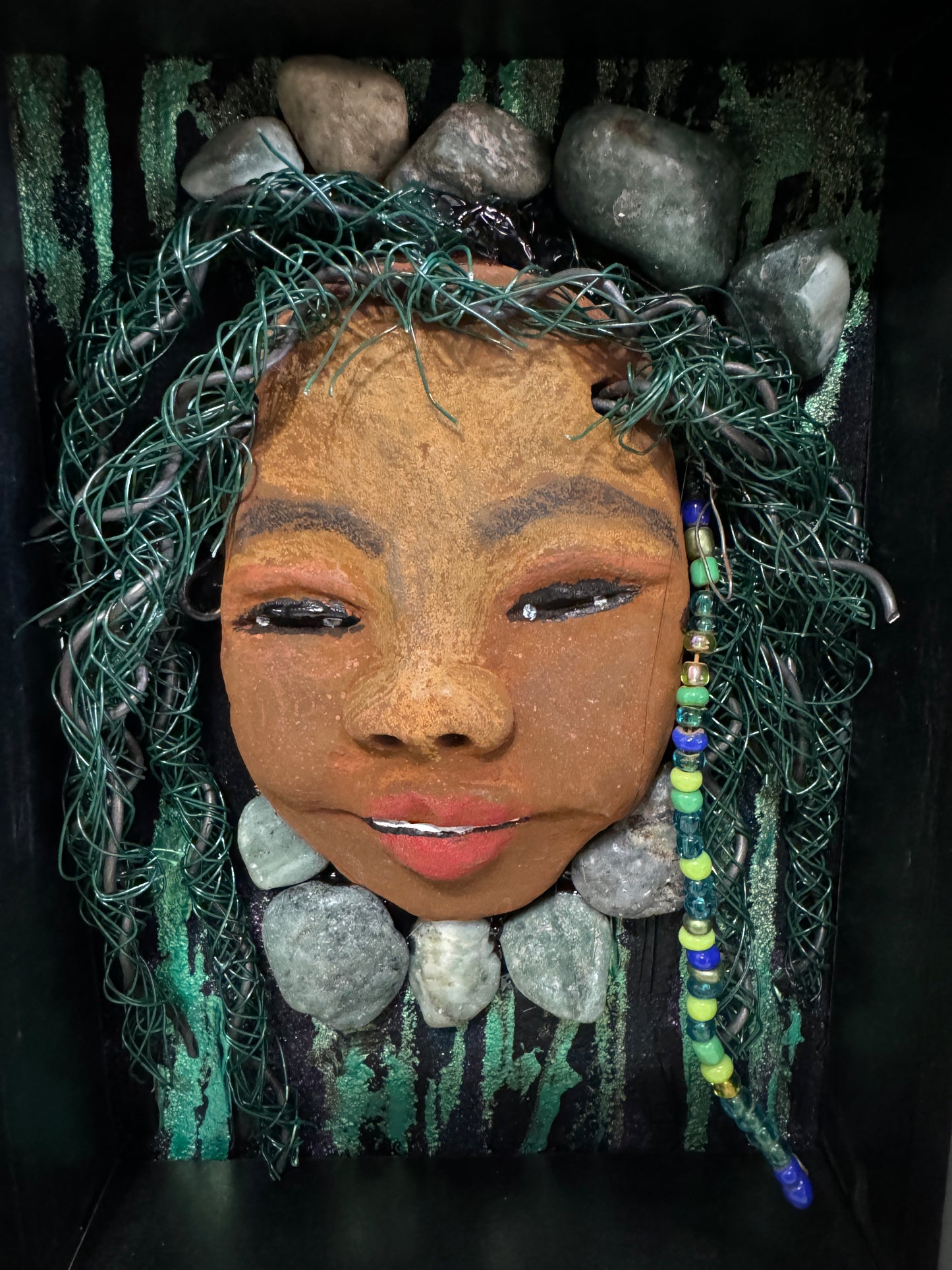 Introducing Allie, a mixed media sculpture raku fired mask. Her features include a two-tone honey brown complexion, Ruby red lips, and hair intricately intertwined with 16 and 24 gauge wire. She is mounted on a hand-painted canvas, accented by  landscaped background, and enclosed in a 5“ x 7“ black shadowbox.