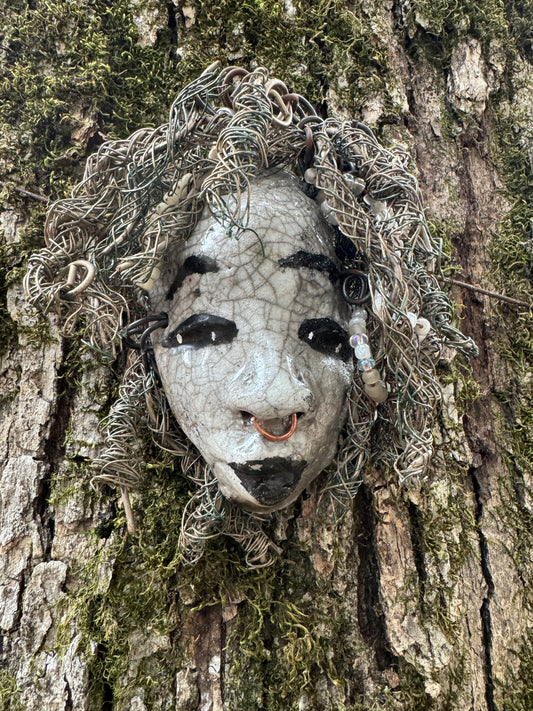 ami is a 4x 6” raku-fired mask featuring a pristine off-white color palette. Her intricately detailed hair is composed of nearly 15 feet of 16 gauge wire, delicately adorned with dainty beads. Set in a backdrop of nature, her portrait makes a wonderful addition to any outdoor art collector, or for decorating the home.