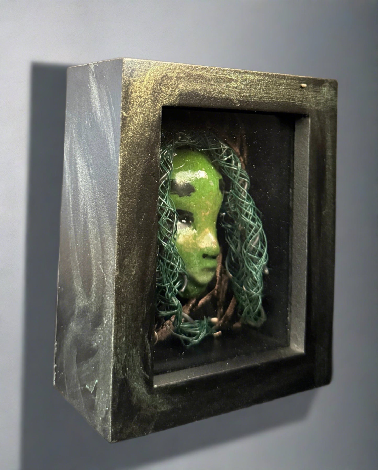 <br>Discover Amoy, a stunning Raku mask made with a blend of materials, housed in a 4" x 5" x 2" antique wood and glass shadowbox.Amoy mesmerizing green appearance and delicately woven hair, crafted from 15 feet of wire and beads, establish it as a unique masterpiece. The hand-painted background depicts Alvita's profound bond with the natural world. With a weight of 10.6 ounces, this expertly crafted mask promises to leave a lasting impression.