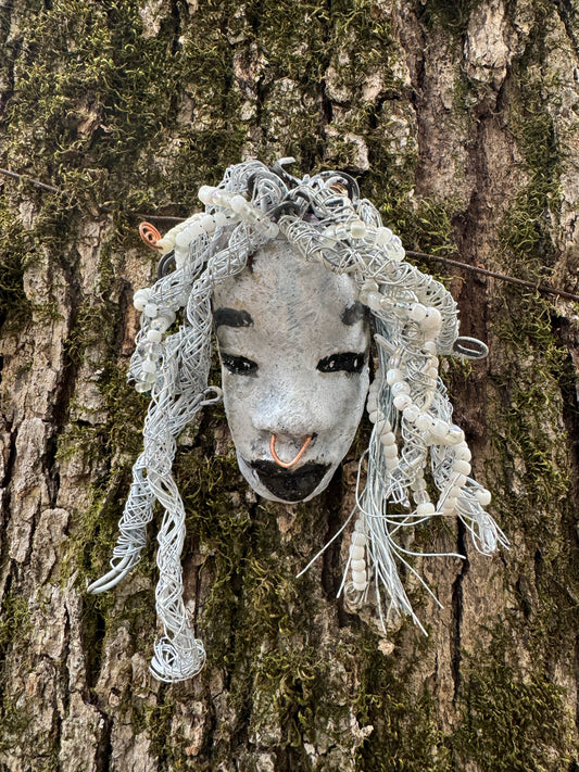 Juanita is a classic 4" x 4" 4.2 oz. Raku-fired mask designed with a white-hued complexion. Her hair is a long 15+ feet of 16-gauge wire, adorned with delicate beads. The portrait of her is set outdoors amongst a tree, making her an ideal present for art and nature admirers, as well as a perfect addition to any outdoor décor.