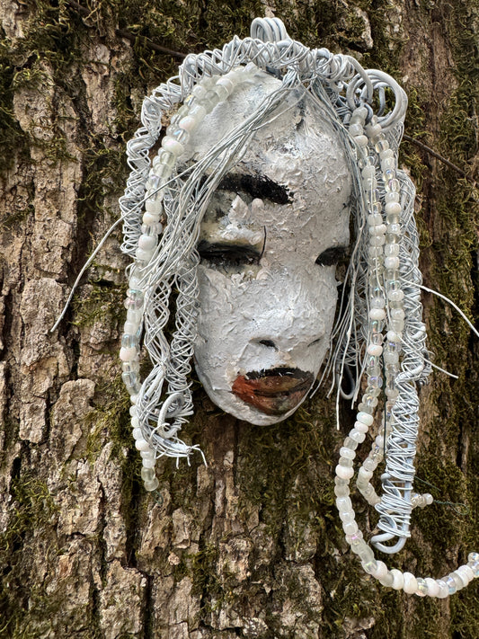 Olivia is a 4 x 7” 5.5 oz raku-fired mask featuring an off-white complexion. 16 gauge wire measuring over 15 feet is used as her hair, accessorized with small decorative beads. Her portrait was taken in an outdoor setting featuring a tree, making her an ideal gift for those who enjoy outdoor art or want to adorn the inside of their home.