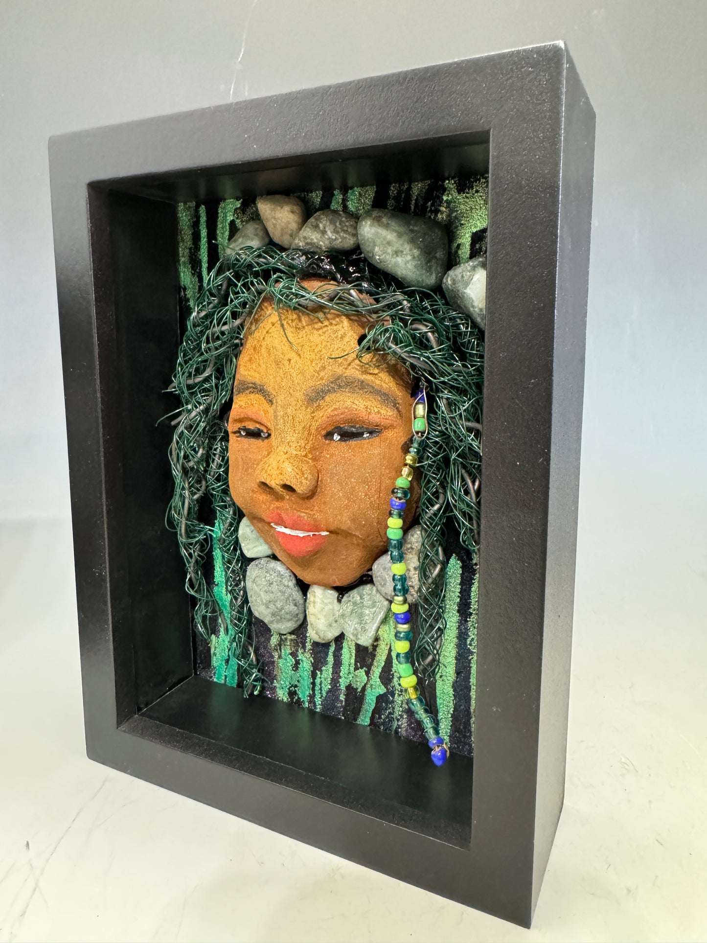 Introducing Allie, a mixed media sculpture raku fired mask. Her features include a two-tone honey brown complexion, Ruby red lips, and hair intricately intertwined with 16 and 24 gauge wire. She is mounted on a hand-painted canvas, accented by  landscaped background, and enclosed in a 5“ x 7“ black shadowbox.