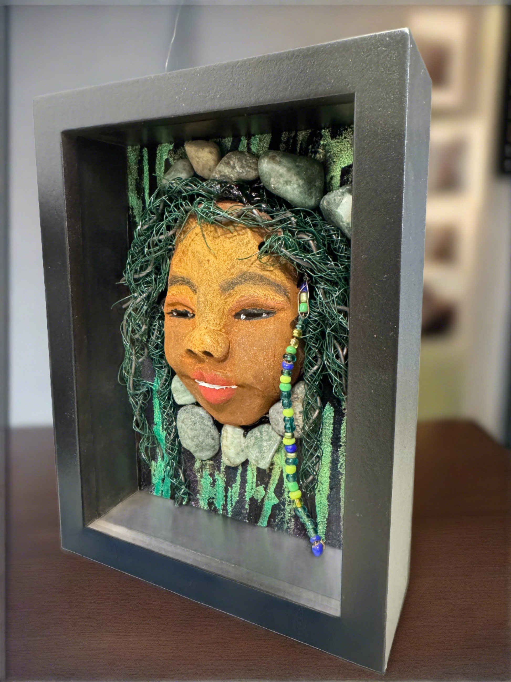 Introducing Allie, a mixed media sculpture raku fired mask. Her features include a two-tone honey brown complexion, Ruby red lips, and hair intricately intertwined with 16 and 24 gauge wire. She is mounted on a hand-painted canvas, accented by landscaped background, and enclosed in a 5“ x 7“ black shadowbox.