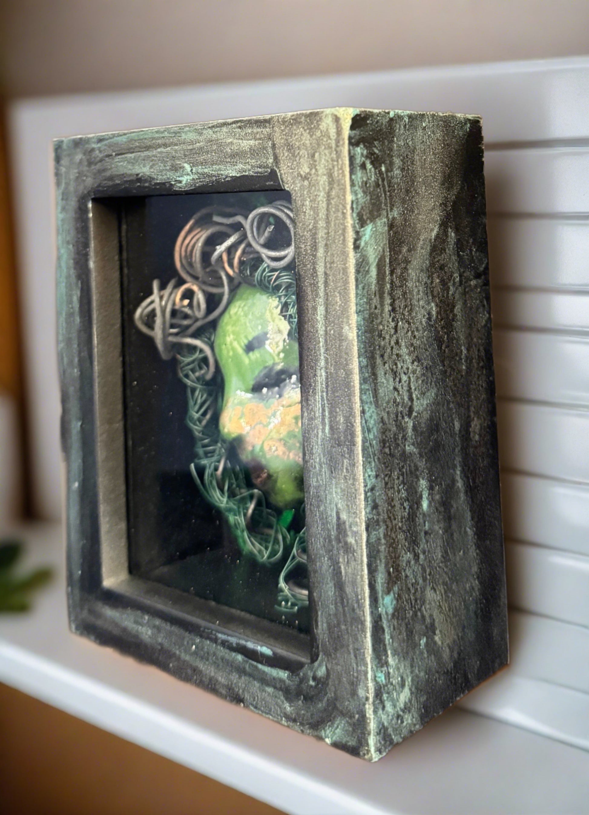 Discover Alvita, a stunning Raku mask made with a blend of materials, housed in a 4" x 5" x 2" antique wood and glass shadowbox. Alvita's mesmerizing green appearance and delicately woven hair, crafted from 15 feet of wire and beads, establish it as a unique masterpiece. The hand-painted background depicts Alvita's profound bond with the natural world. With a weight of 10.6 ounces, this expertly crafted mask promises to leave a lasting impression.