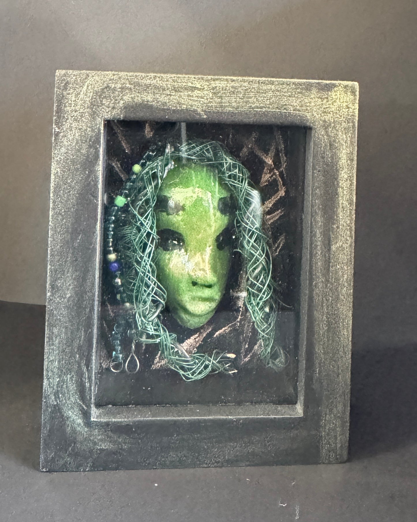 <br>Discover Amoy, a stunning Raku mask made with a blend of materials, housed in a 4" x 5" x 2" antique wood and glass shadowbox.Amoy mesmerizing green appearance and delicately woven hair, crafted from 15 feet of wire and beads, establish it as a unique masterpiece. The hand-painted background depicts Alvita's profound bond with the natural world. With a weight of 10.6 ounces, this expertly crafted mask promises to leave a lasting impression.