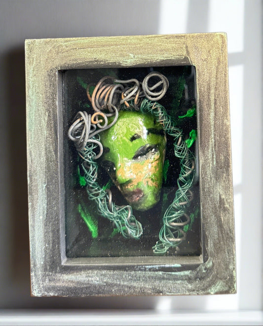 Discover Alvita, a stunning Raku mask made with a blend of materials, housed in a 4" x 5" x 2" antique wood and glass shadowbox. Alvita's mesmerizing green appearance and delicately woven hair, crafted from 15 feet of wire and beads, establish it as a unique masterpiece. The hand-painted background depicts Alvita's profound bond with the natural world. With a weight of 10.6 ounces, this expertly crafted mask promises to leave a lasting impression.