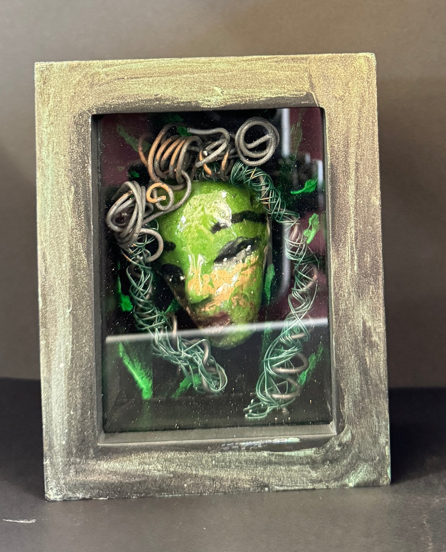 Discover Alvita, a stunning Raku mask made with a blend of materials, housed in a 4" x 5" x 2" antique wood and glass shadowbox. Alvita's mesmerizing green appearance and delicately woven hair, crafted from 15 feet of wire and beads, establish it as a unique masterpiece. The hand-painted background depicts Alvita's profound bond with the natural world. With a weight of 10.6 ounces, this expertly crafted mask promises to leave a lasting impression.