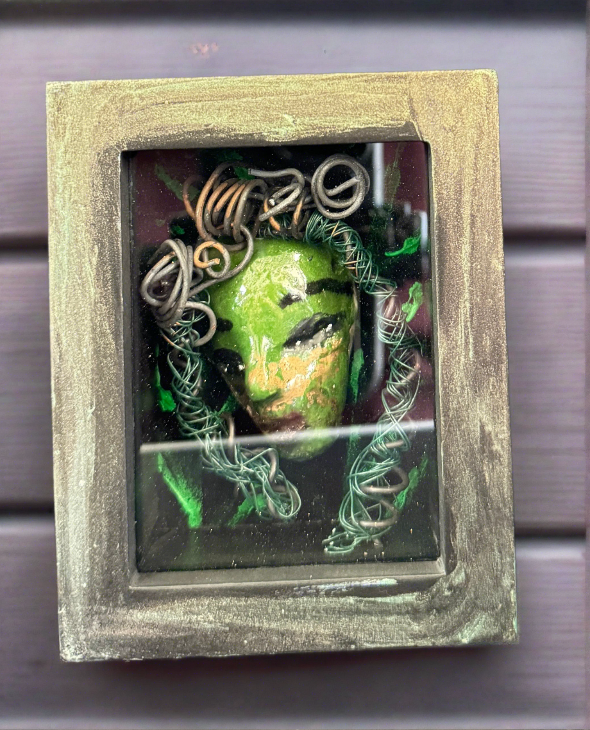 Discover Alvita, a stunning Raku mask made with a blend of materials, housed in a 4" x 5" x 2" antique wood and glass shadowbox. Alvita's mesmerizing green appearance and delicately woven hair, crafted from 15 feet of wire and beads, establish it as a unique masterpiece. The hand-painted background depicts Alvita's profound bond with the natural world. With a weight of 10.6 ounces, this expertly crafted mask promises to leave a lasting impression.