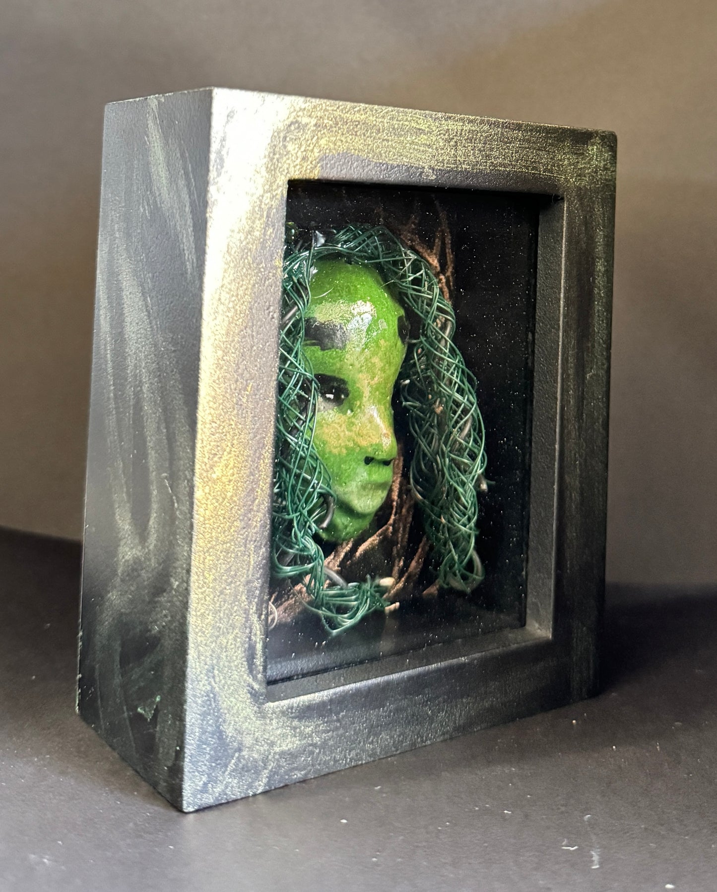 <br>Discover Amoy, a stunning Raku mask made with a blend of materials, housed in a 4" x 5" x 2" antique wood and glass shadowbox.Amoy mesmerizing green appearance and delicately woven hair, crafted from 15 feet of wire and beads, establish it as a unique masterpiece. The hand-painted background depicts Alvita's profound bond with the natural world. With a weight of 10.6 ounces, this expertly crafted mask promises to leave a lasting impression.