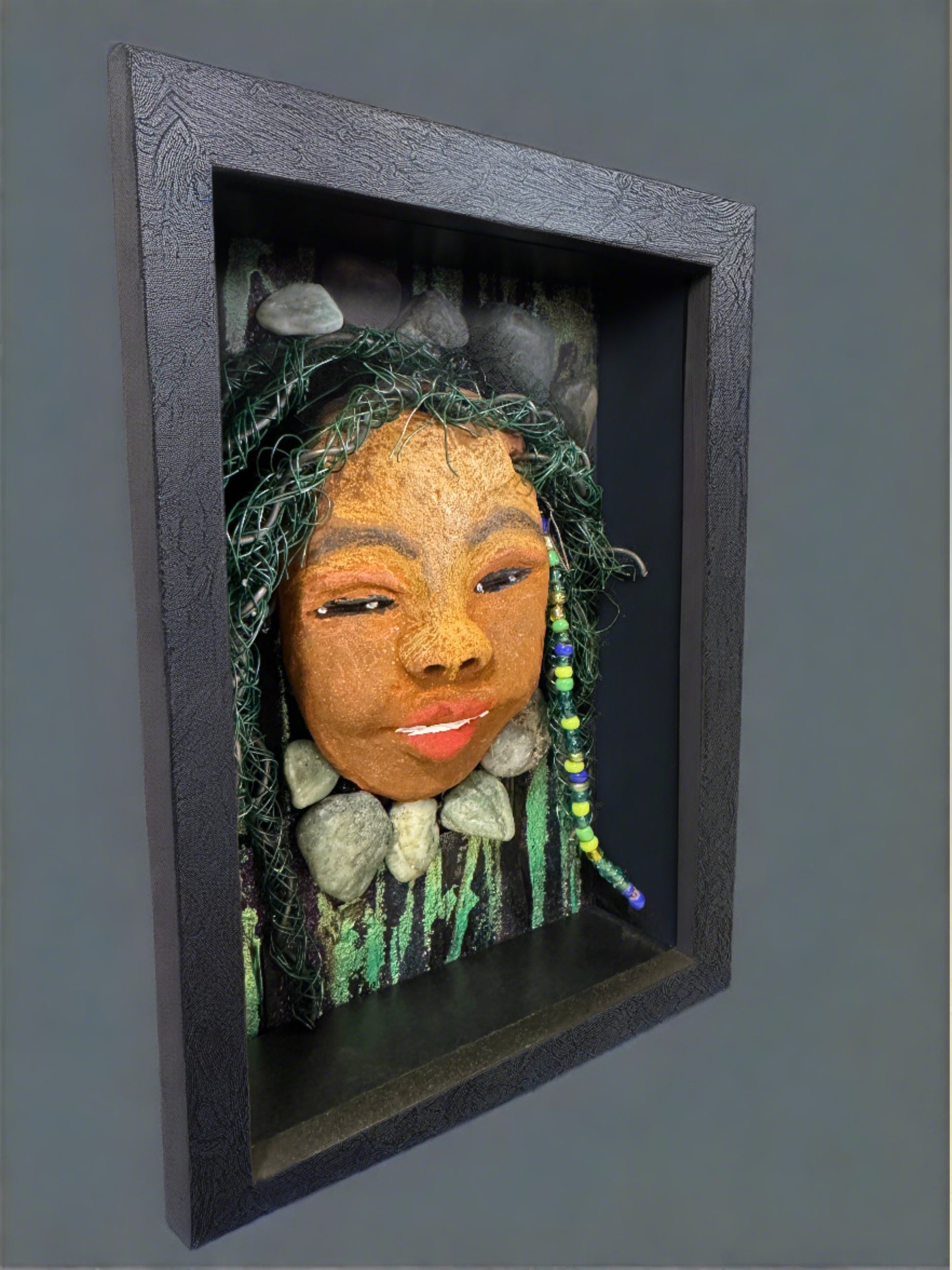 Introducing Allie, a mixed media sculpture raku fired mask. Her features include a two-tone honey brown complexion, Ruby red lips, and hair intricately intertwined with 16 and 24 gauge wire. She is mounted on a hand-painted canvas, accented by  landscaped background, and enclosed in a 5“ x 7“ black shadowbox.