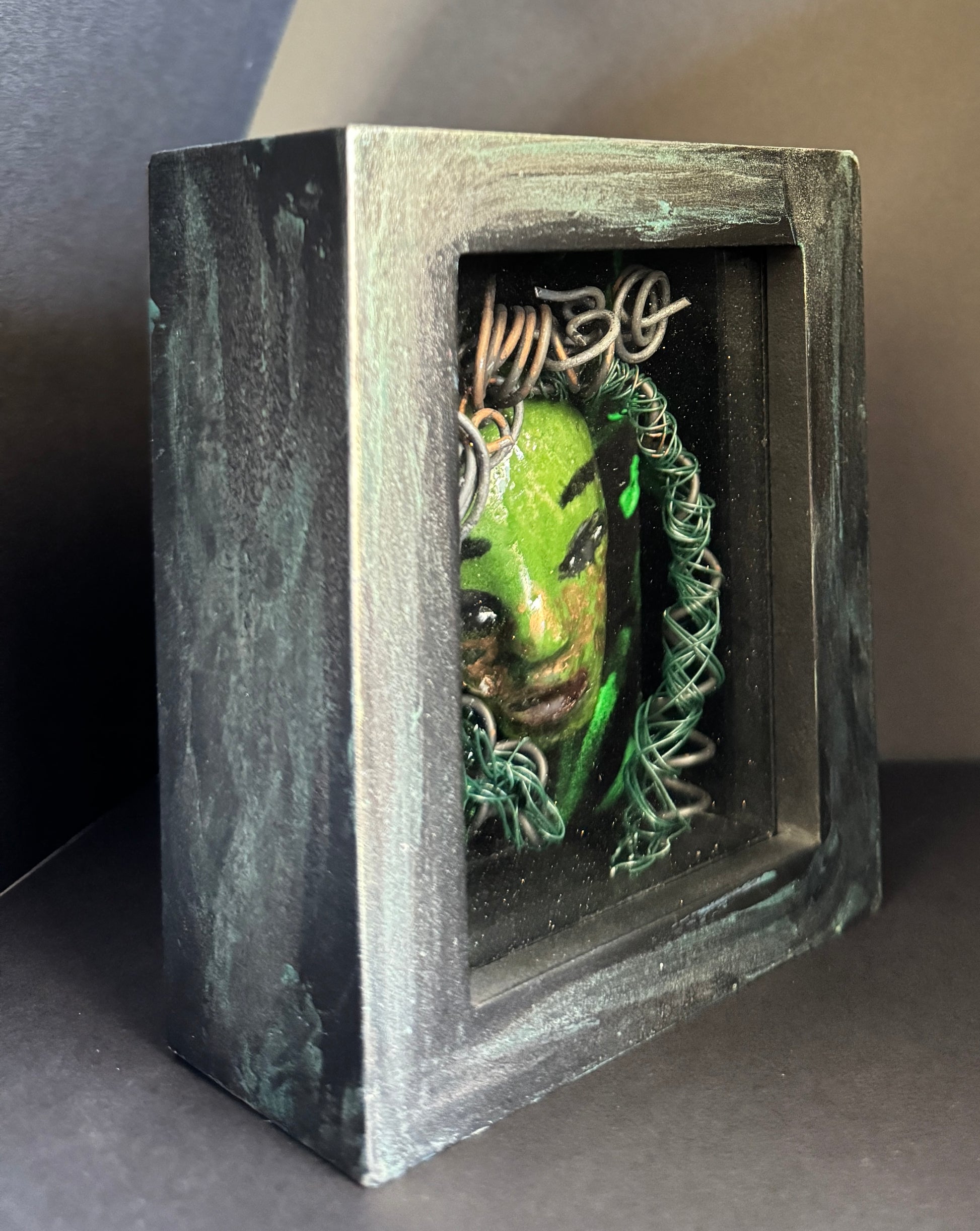 Discover Alvita, a stunning Raku mask made with a blend of materials, housed in a 4" x 5" x 2" antique wood and glass shadowbox. Alvita's mesmerizing green appearance and delicately woven hair, crafted from 15 feet of wire and beads, establish it as a unique masterpiece. The hand-painted background depicts Alvita's profound bond with the natural world. With a weight of 10.6 ounces, this expertly crafted mask promises to leave a lasting impression.