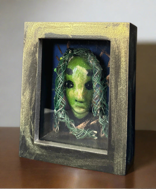 <br>Discover Amoy, a stunning Raku mask made with a blend of materials, housed in a 4" x 5" x 2" antique wood and glass shadowbox.Amoy mesmerizing green appearance and delicately woven hair, crafted from 15 feet of wire and beads, establish it as a unique masterpiece. The hand-painted background depicts Alvita's profound bond with the natural world. With a weight of 10.6 ounces, this expertly crafted mask promises to leave a lasting impression.