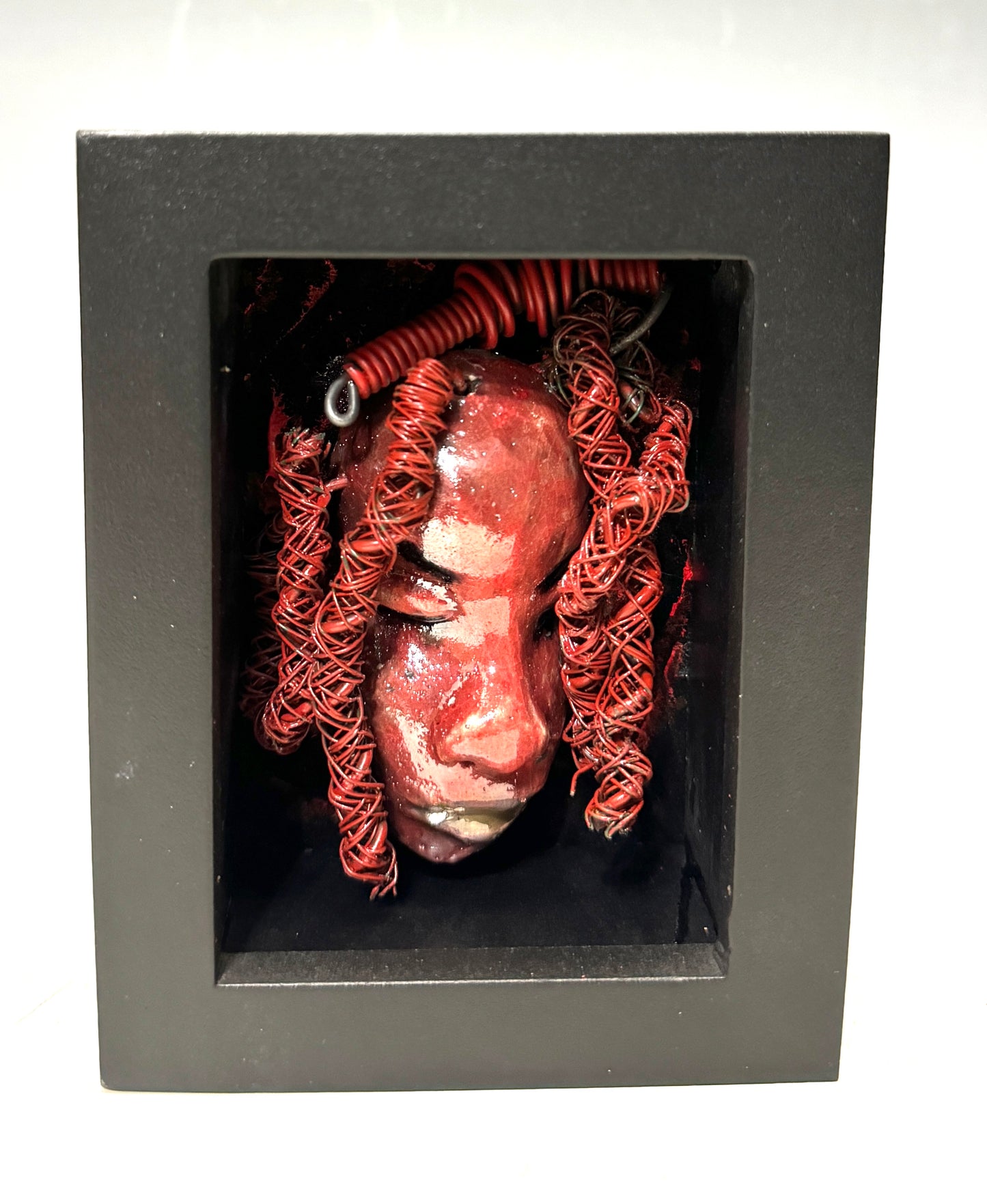 Discover your daring side with Rose - a striking, one-of-a-kind mixed media mask that has been raku fired and placed in a 4" x 4" black shadowbox. Featuring over 25 feet of 16 and 24 gauge wire as hair, this bold and unique piece will add a touch of adventure to any space. Embrace the unconventional and stand out from the ordinary with Rose, the statement piece that will elevate your decor with its fiery presence.