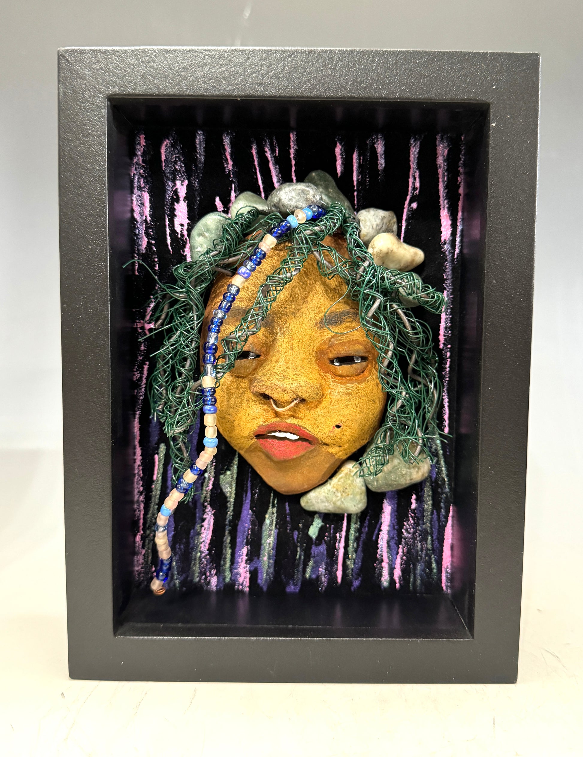 Introducing Beverly: A striking mixed media mask created using the raku firing method. The meticulously designed features showcase a beautiful honey brown complexion with subtle Ruby red lips and intricately woven hair made with 16 and 24 gauge wire. This exquisite piece is mounted on a hand-painted canvas, featuring a stunningly landscaped background, and elegantly presented in a glass covered 5" x 7" black shadowbox.